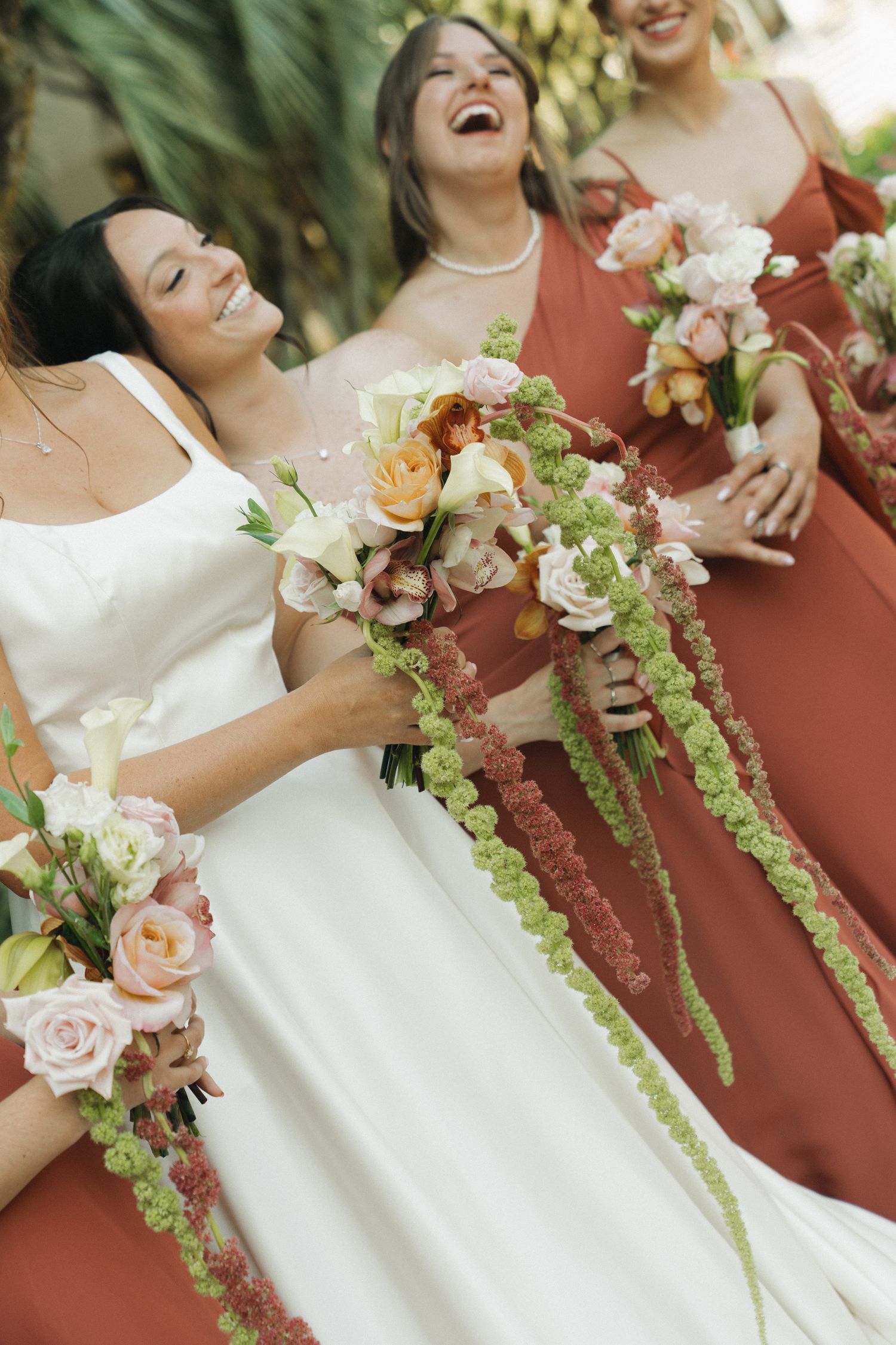 Bride in white gown laughing with bridesmaids in rust dresses holding cascading floral bouquets.