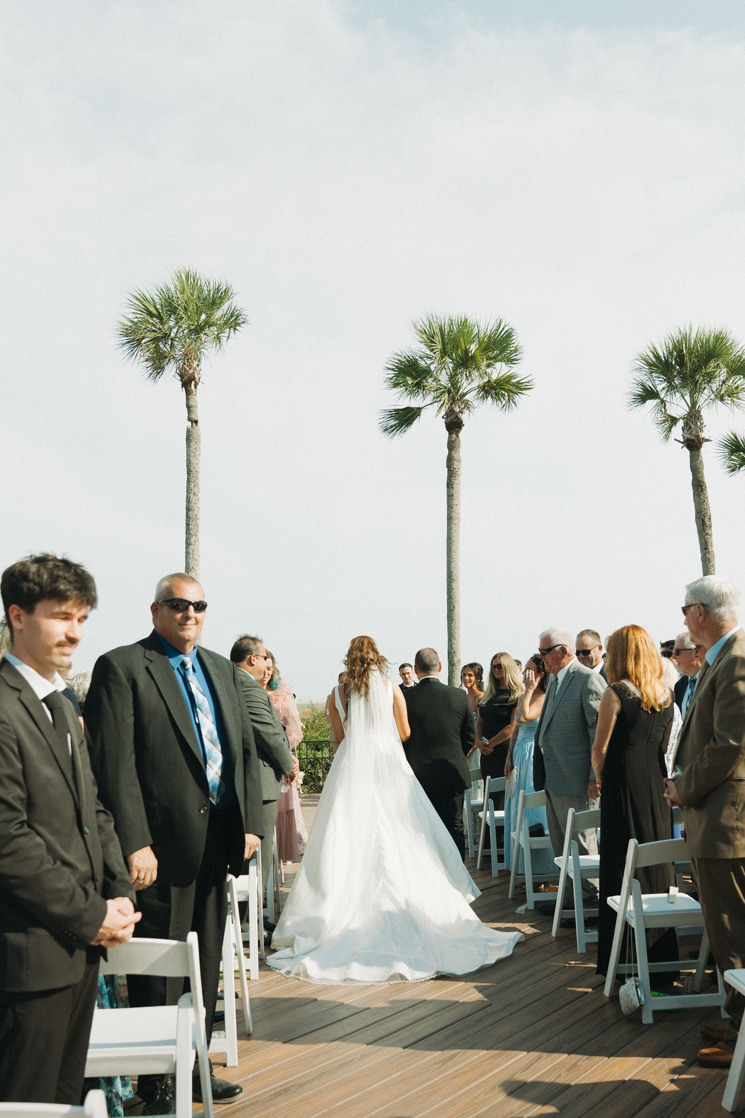 Bride walks down the aisle at an outdoor wedding ceremony on a deck with palm trees and white chairs.