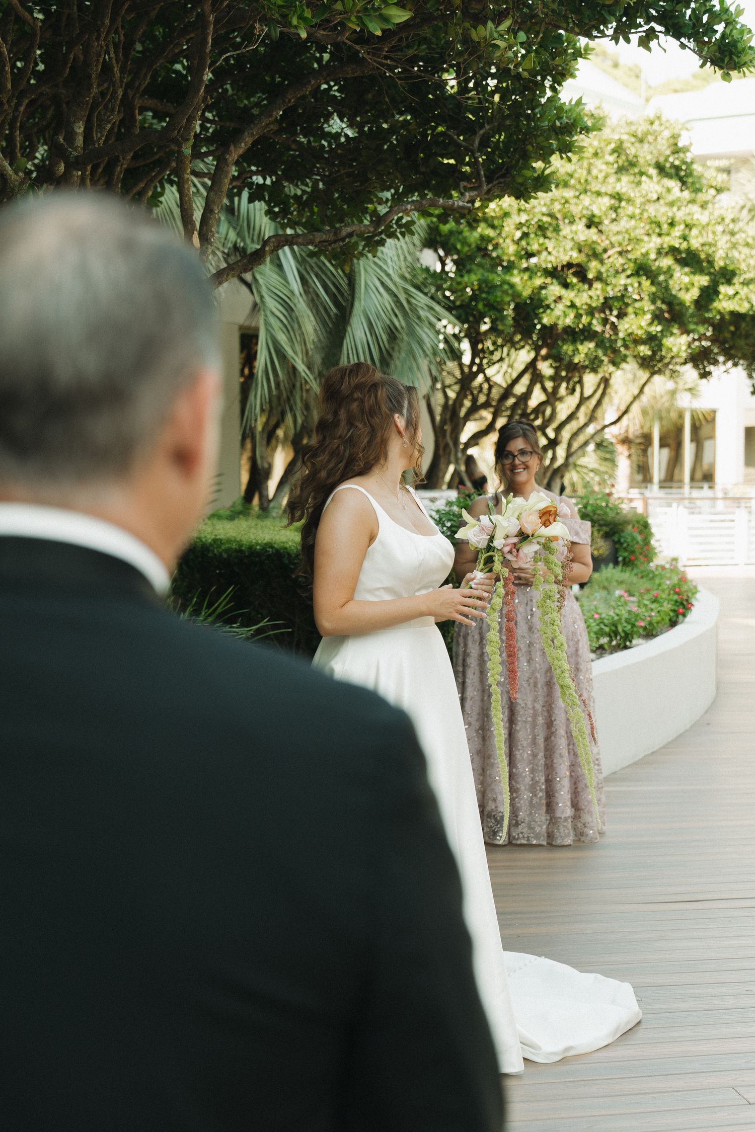 Bride in white gown holds bouquet outdoors with bridesmaid in floral dress, as groom looks on at garden wedding.