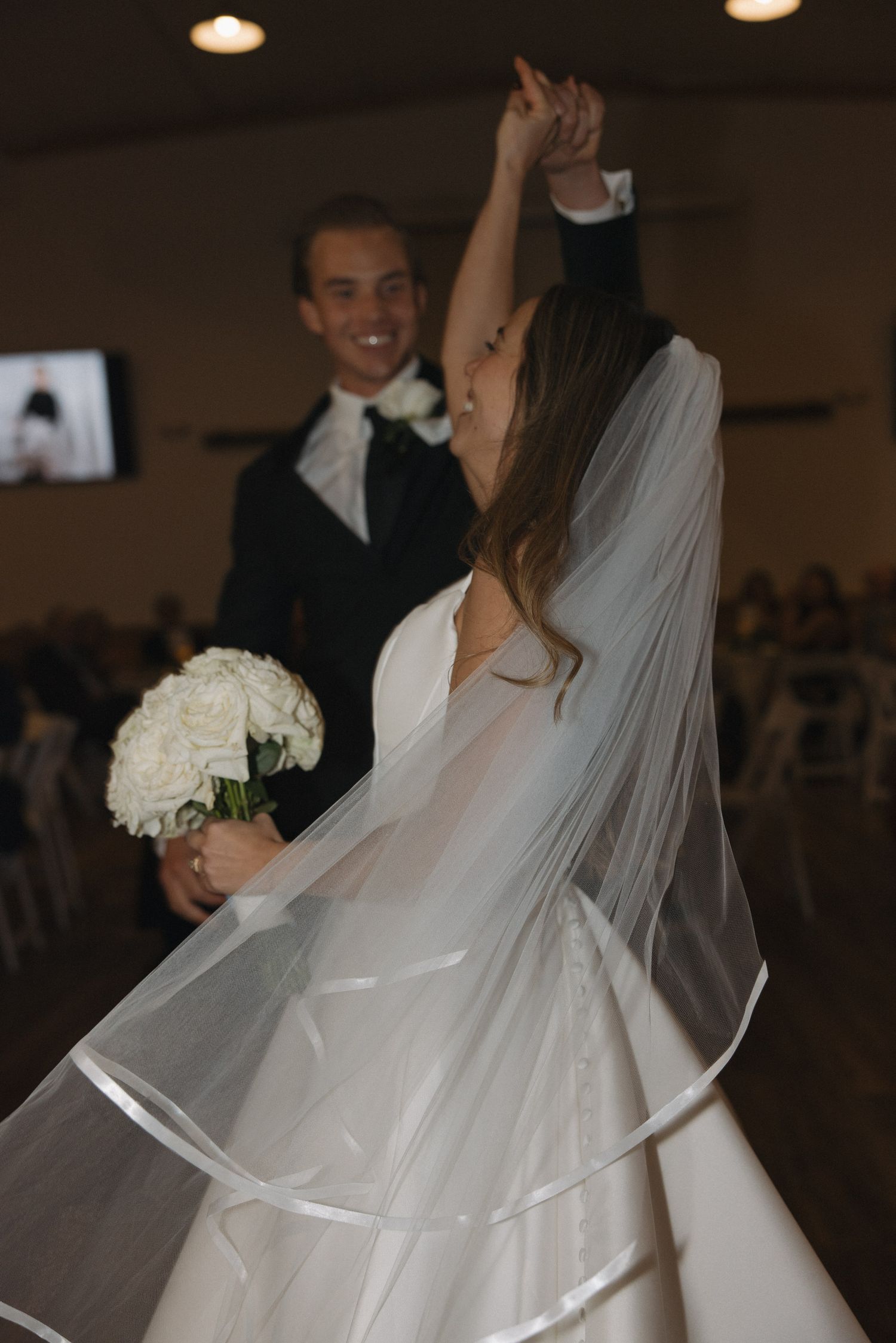 Bride in white gown and veil twirls with bouquet as groom in black suit smiles during wedding reception dance.