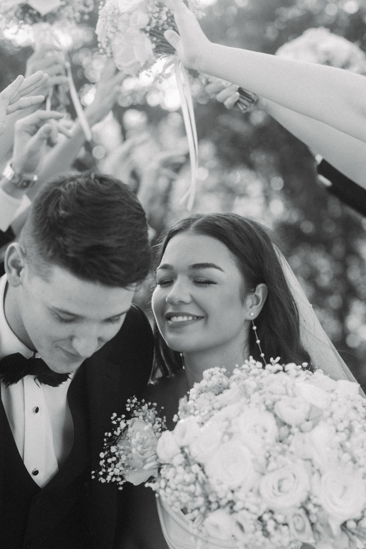 Black and white photo of smiling bride and groom surrounded by guests raising flowers at wedding ceremony.