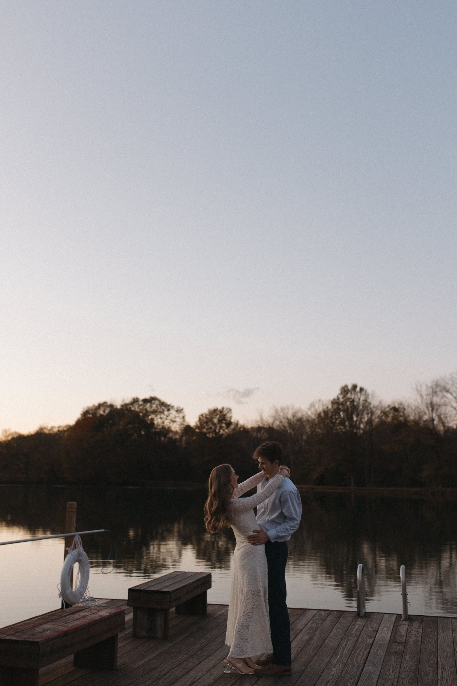 Couple embracing on a wooden dock by a calm lake during a peaceful autumn sunset.