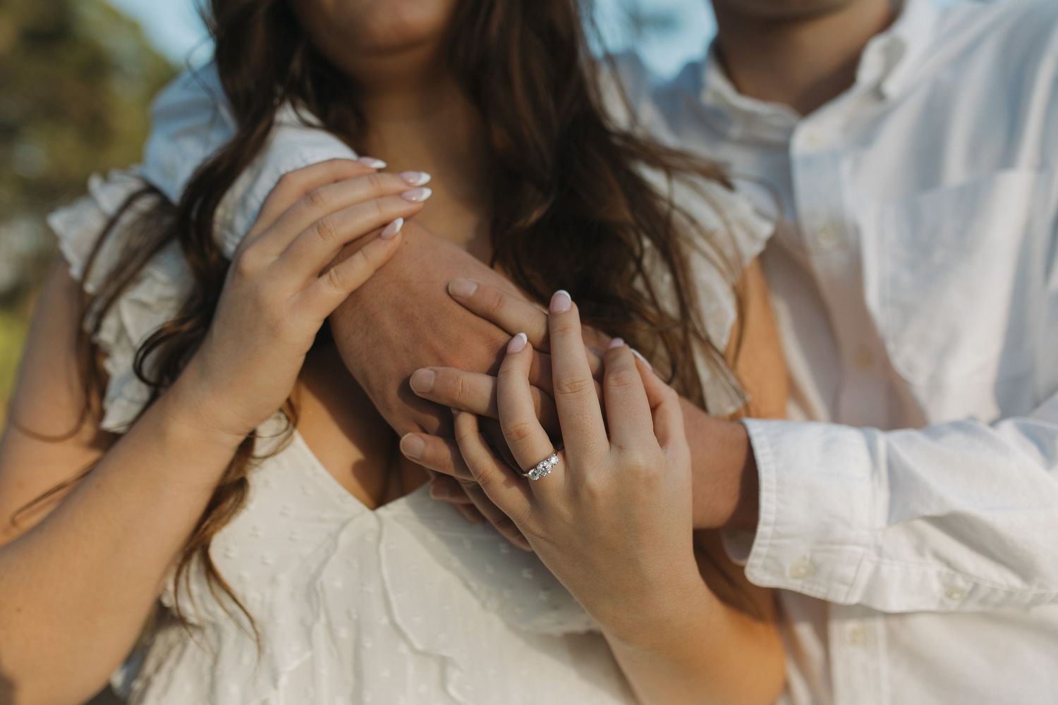 Close-up of a couple holding hands, highlighting a diamond engagement ring outdoors.