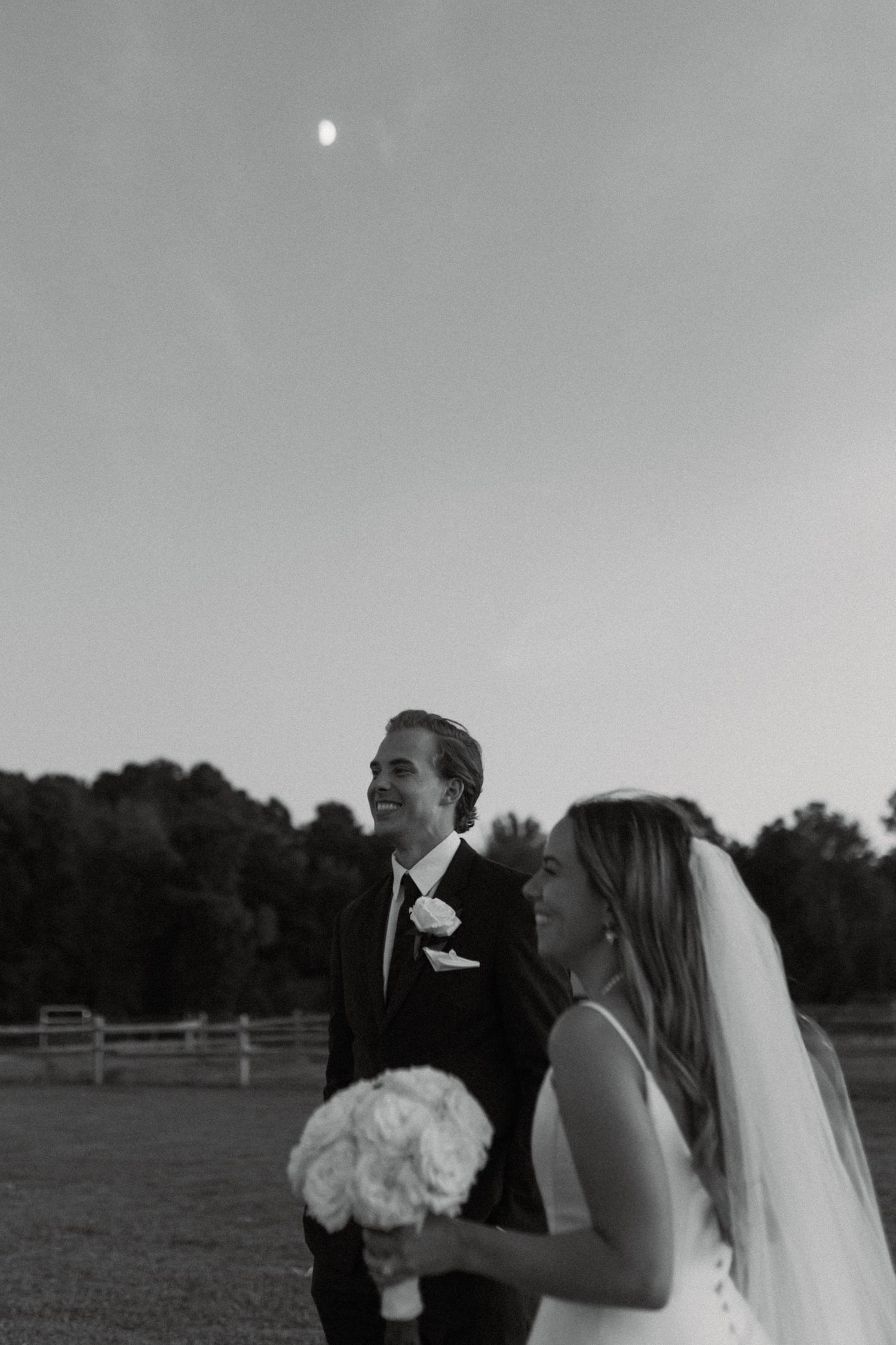 Black and white photo of smiling bride and groom outdoors at dusk, bride holding white bouquet.