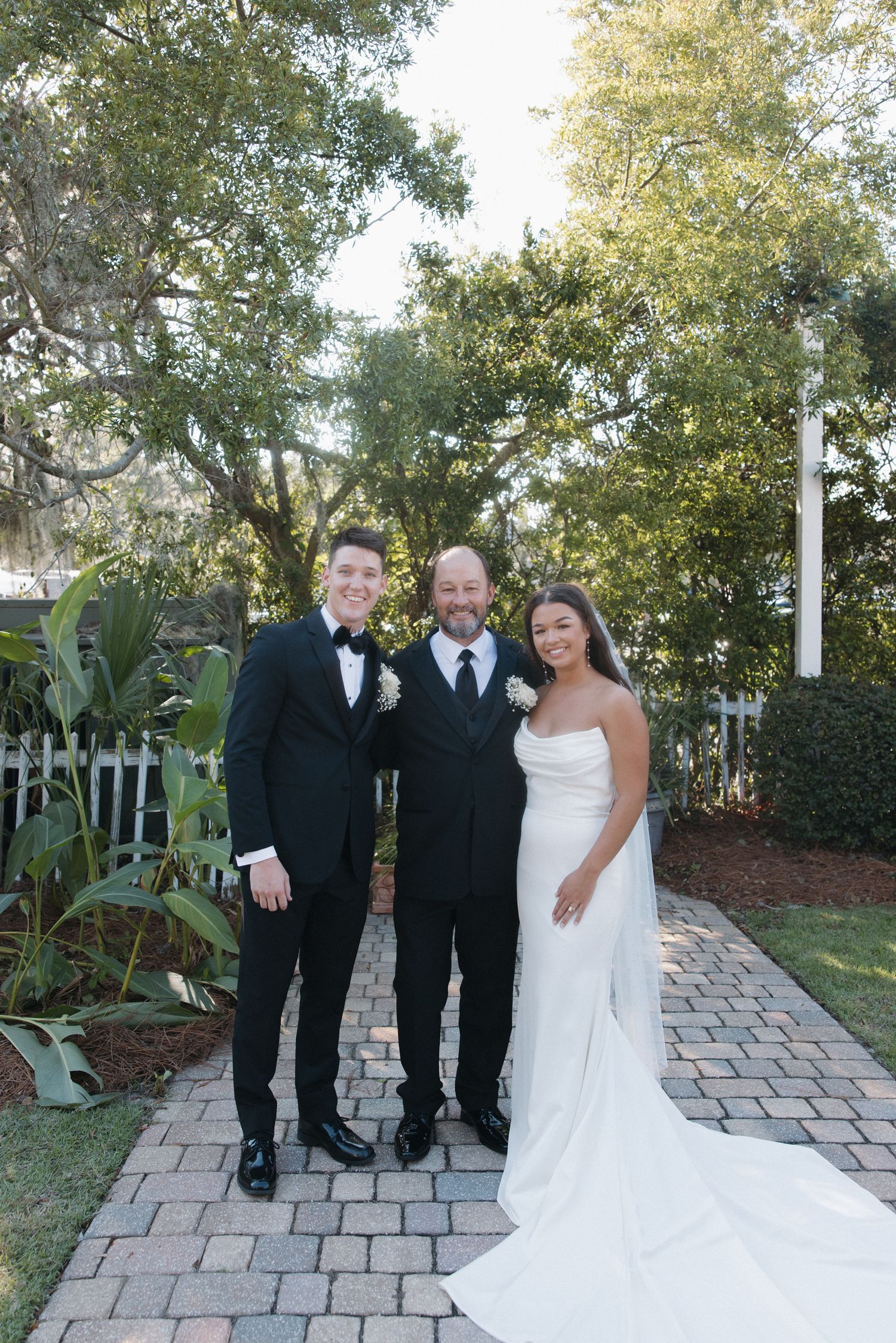 Bride in white gown and groom in tuxedo pose with officiant on brick path surrounded by lush greenery.
