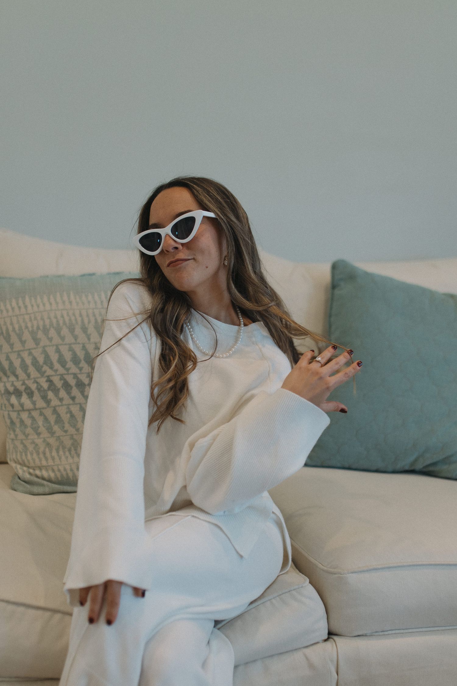 Woman in white outfit and cat-eye sunglasses sitting on a cream sofa with teal pillows.