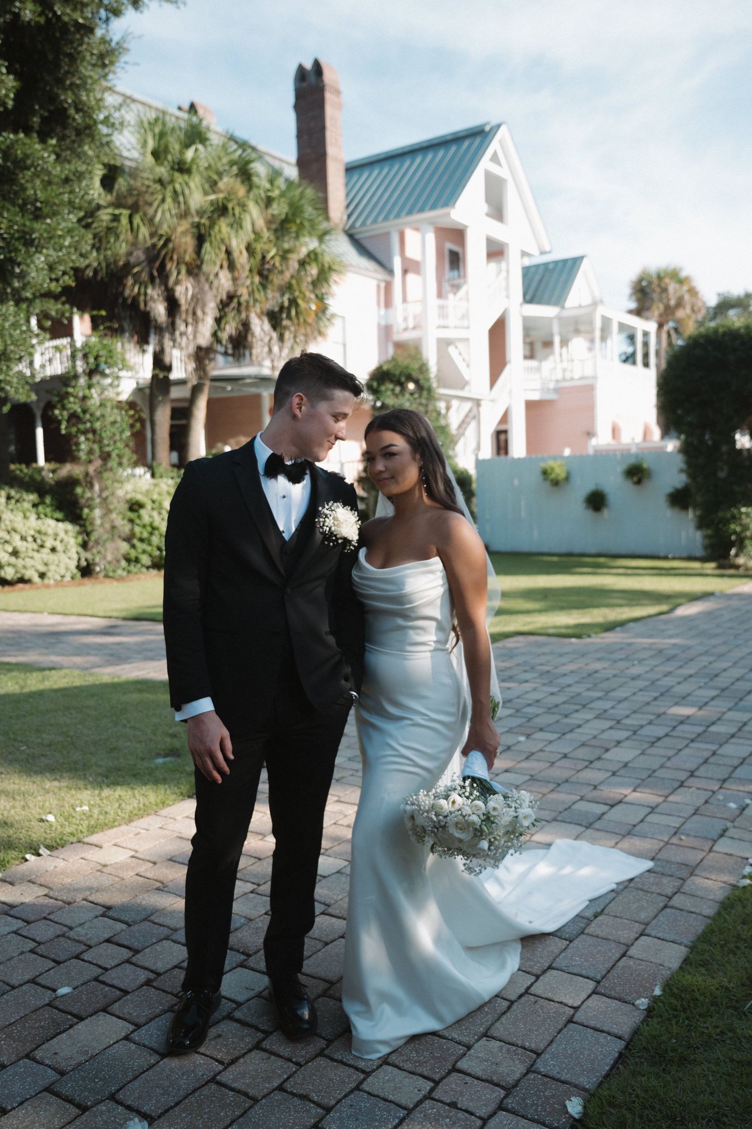 Bride in sleek white gown and groom in black tuxedo walking together on brick path at outdoor wedding venue.