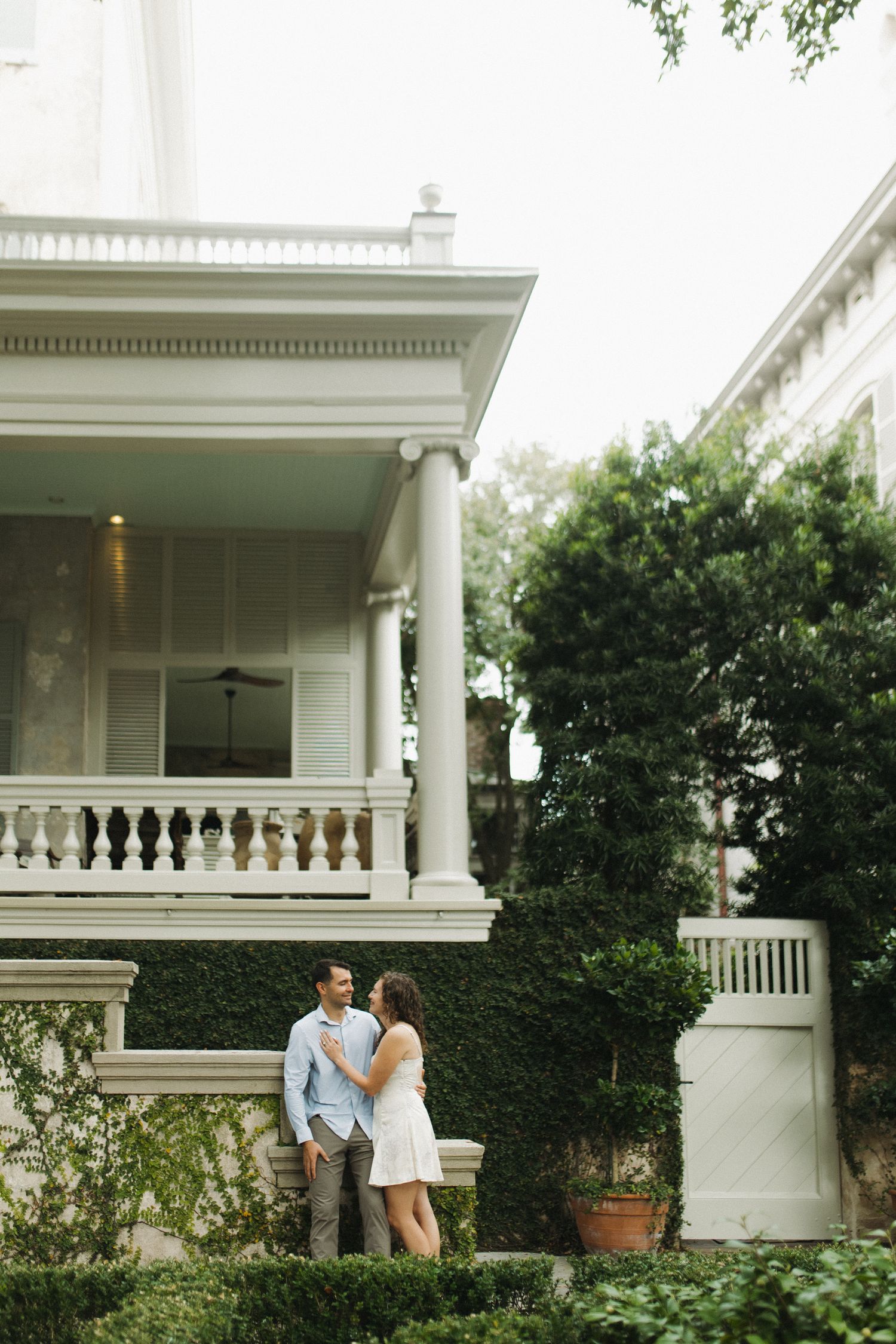 Couple standing together in front of an elegant white Southern mansion with balcony and lush greenery.