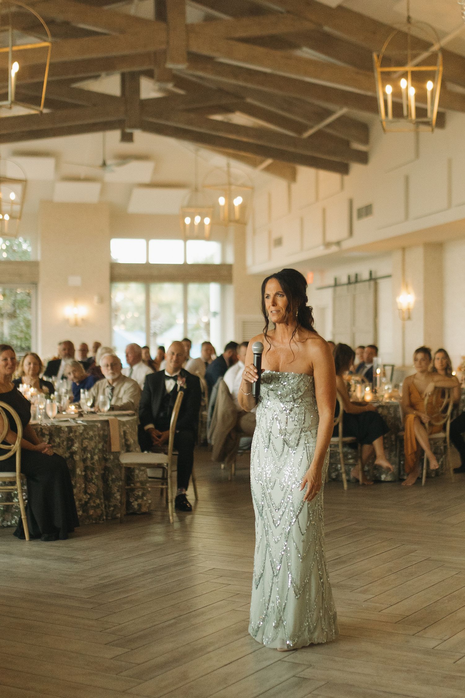 Woman in beaded strapless gown giving a speech at an elegant wedding reception with guests seated at decorated tables.