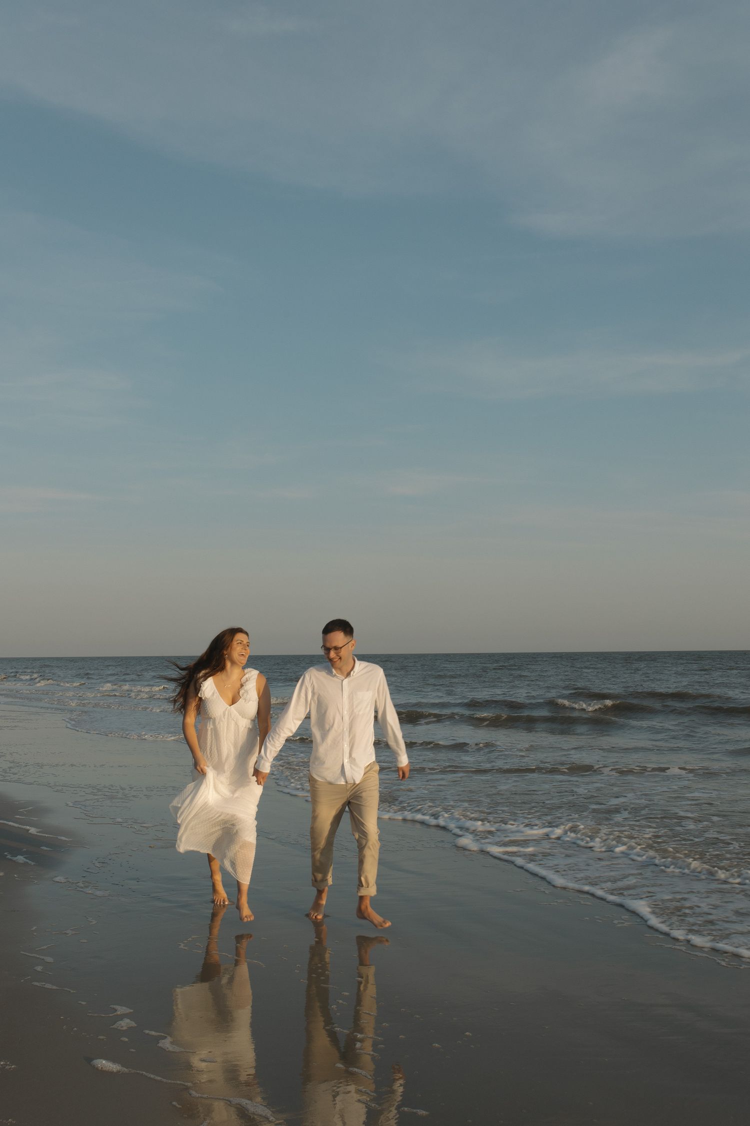 Couple walks barefoot along a reflective shoreline at sunset in white outfits.