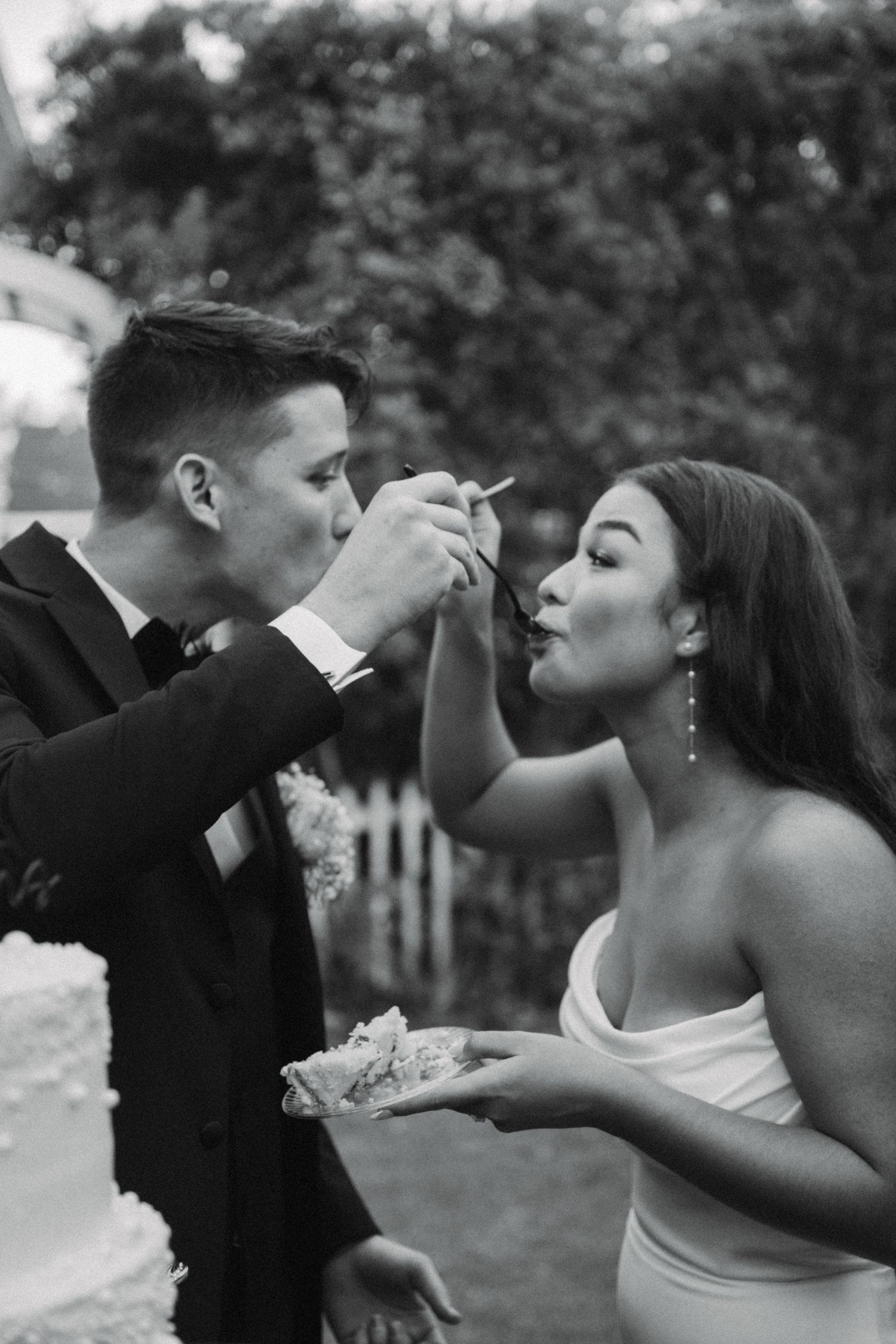 Bride and groom share wedding cake during intimate black and white outdoor reception moment.