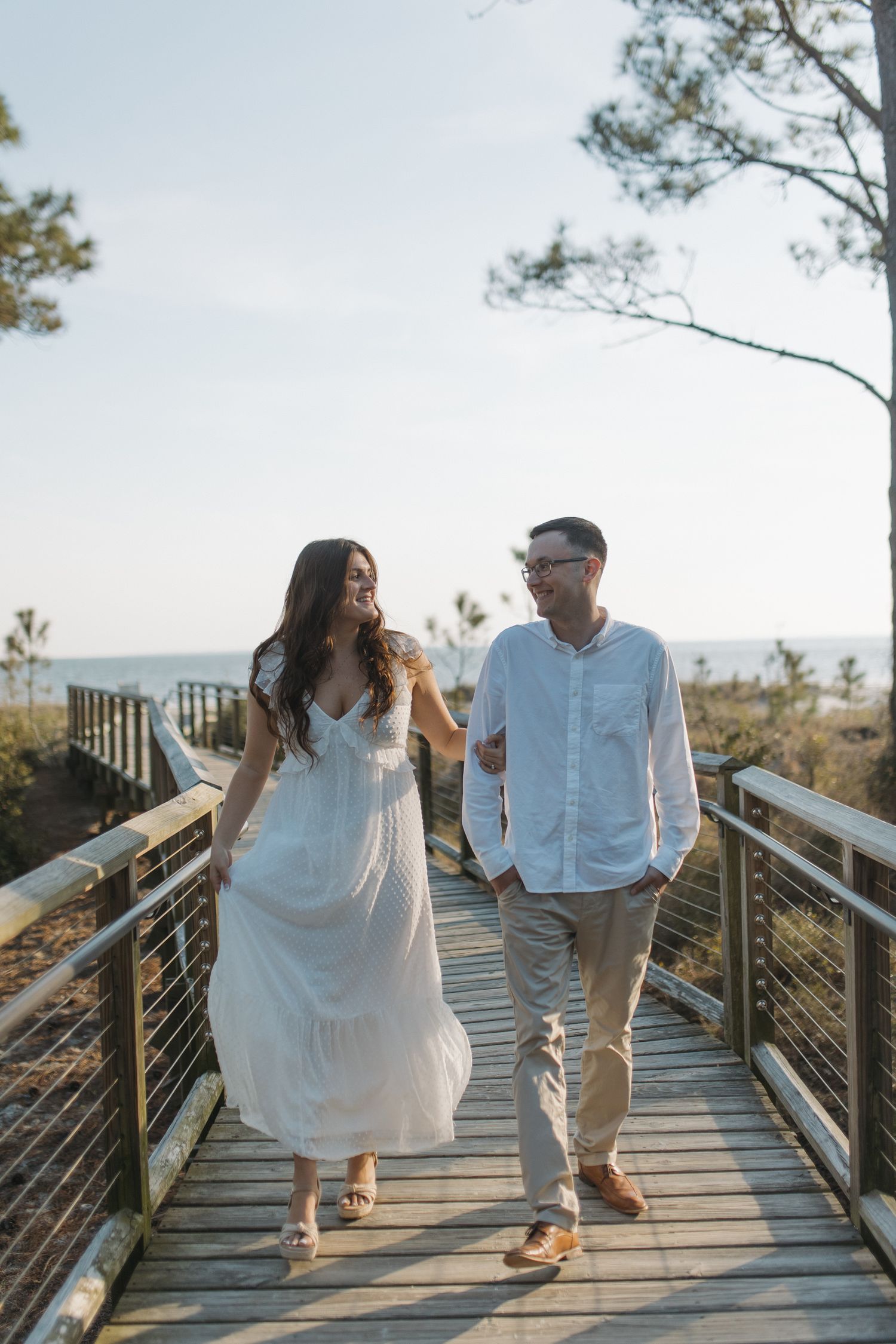 Couple walks hand in hand along a wooden boardwalk overlooking the ocean.