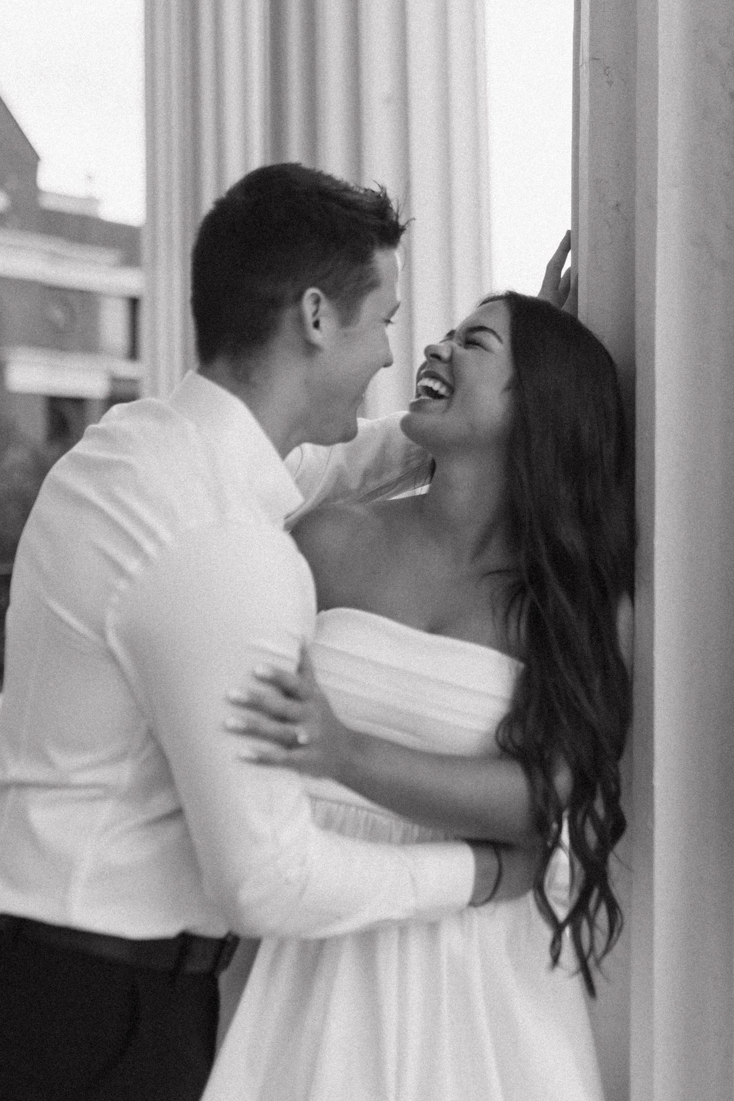 Black and white photo of couple laughing and embracing against a white column outdoors.