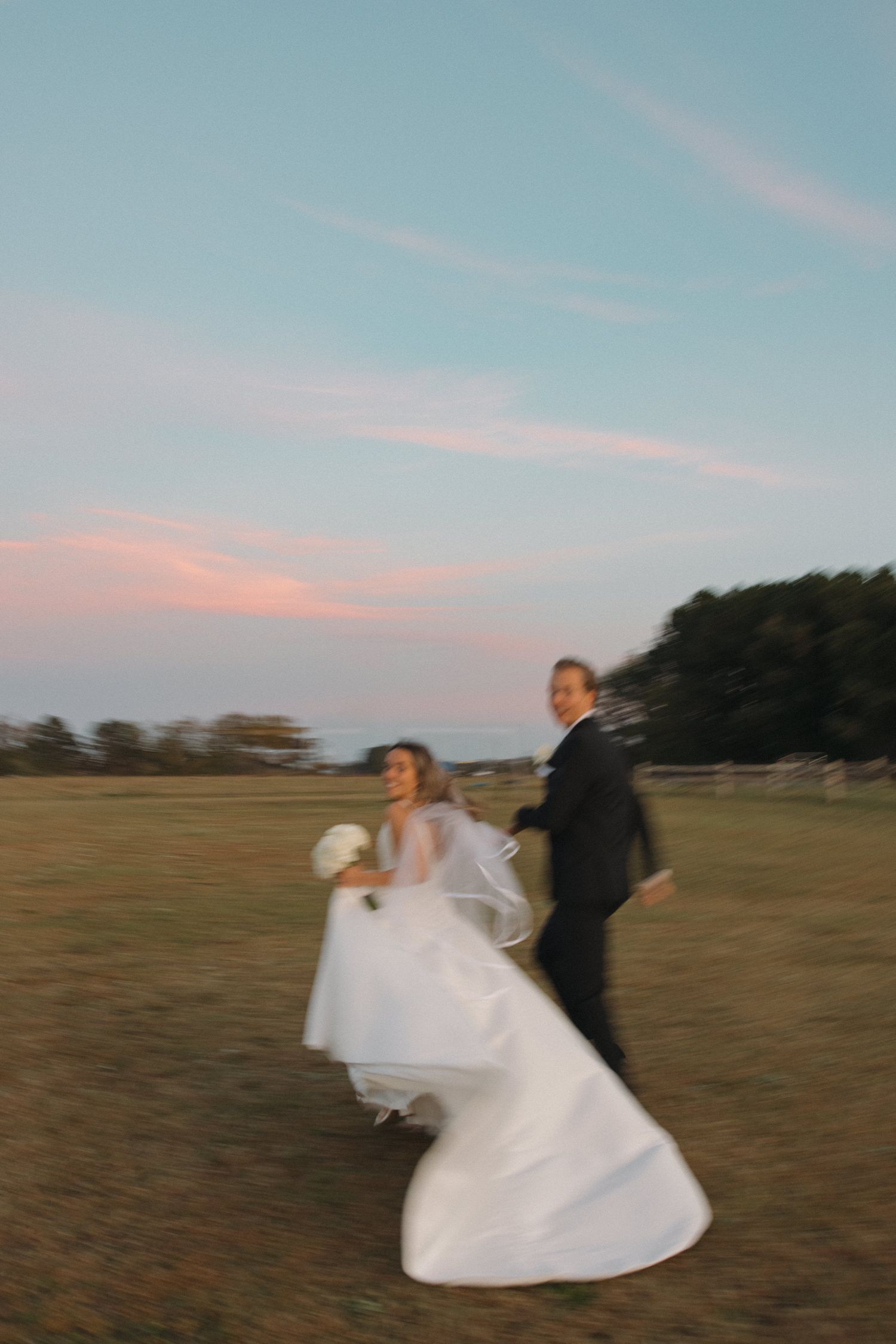 Bride and groom run joyfully across an open field at dusk, wedding dress flowing in motion under a pastel sky.