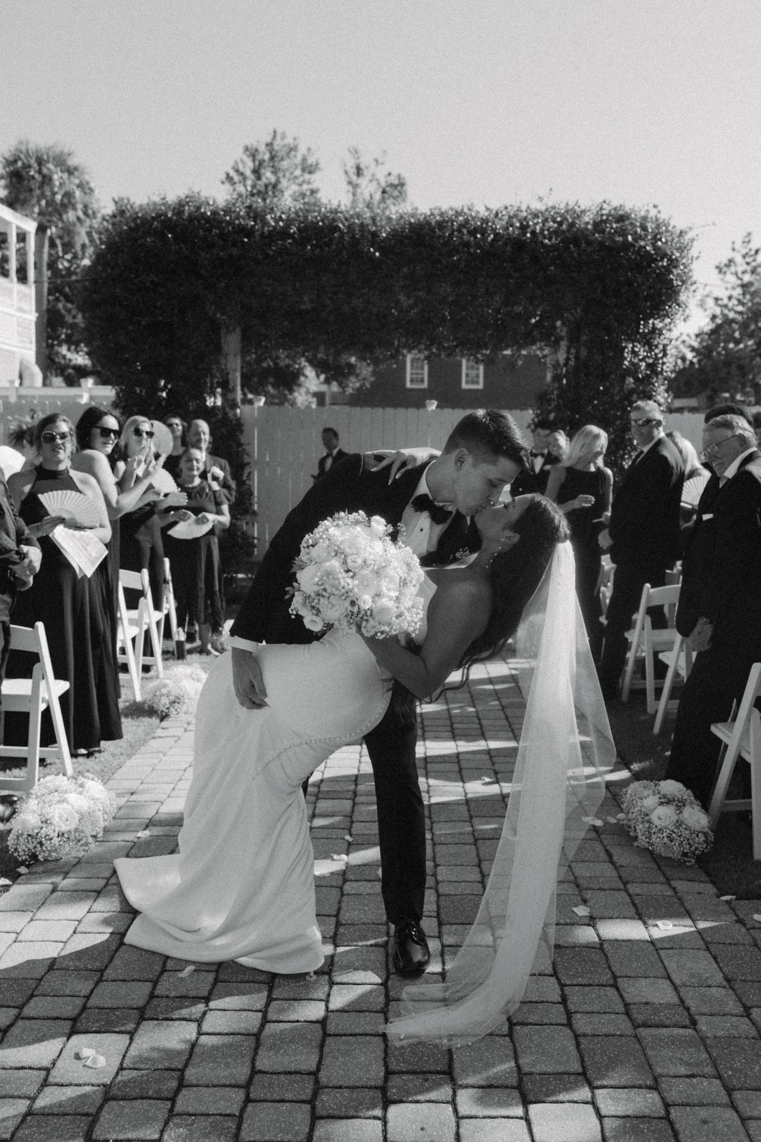 Black and white photo of groom dipping bride for kiss at outdoor wedding ceremony with guests seated alongside.