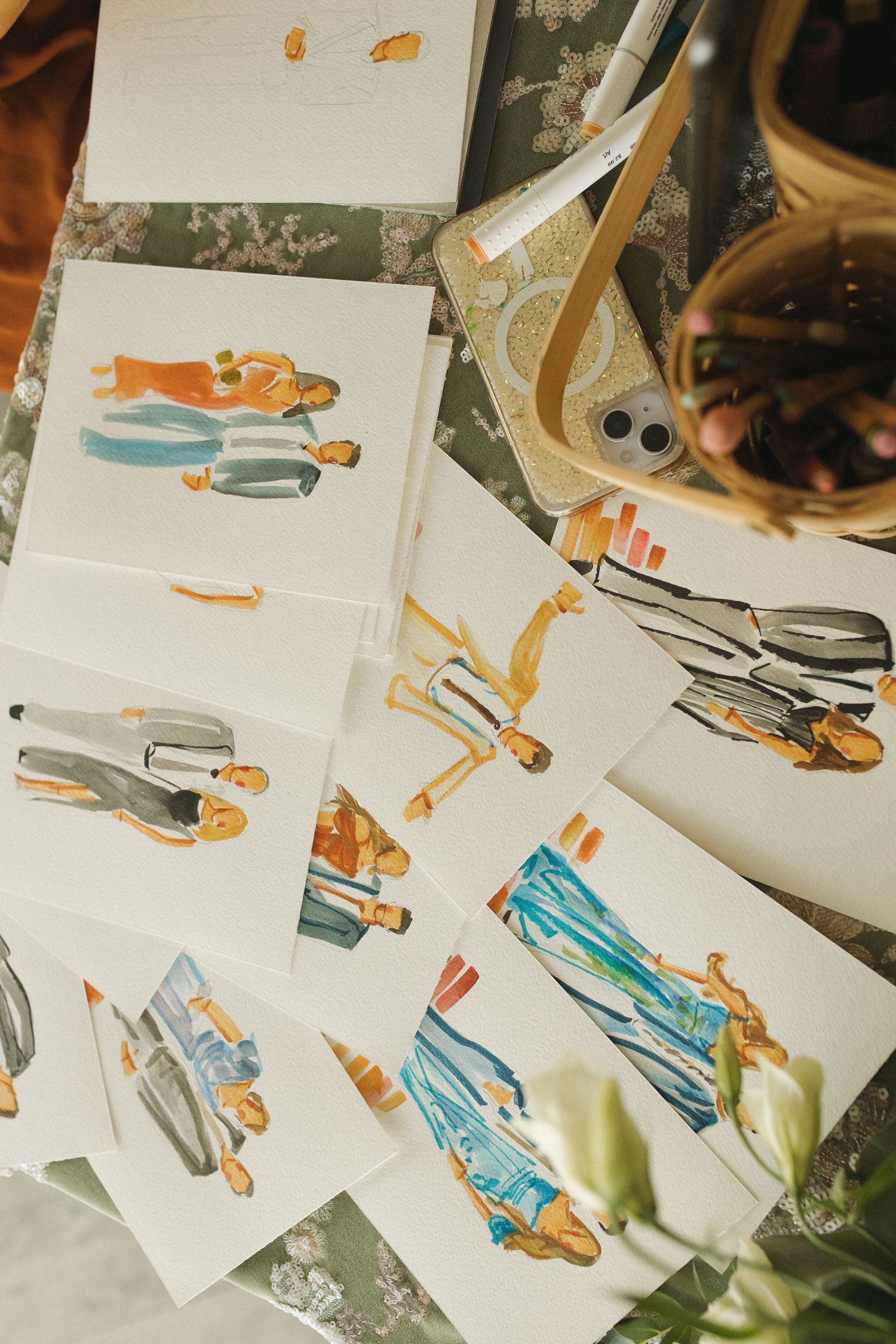 Fashion illustration sketches of figures in colorful outfits spread across a floral table with art supplies nearby.
