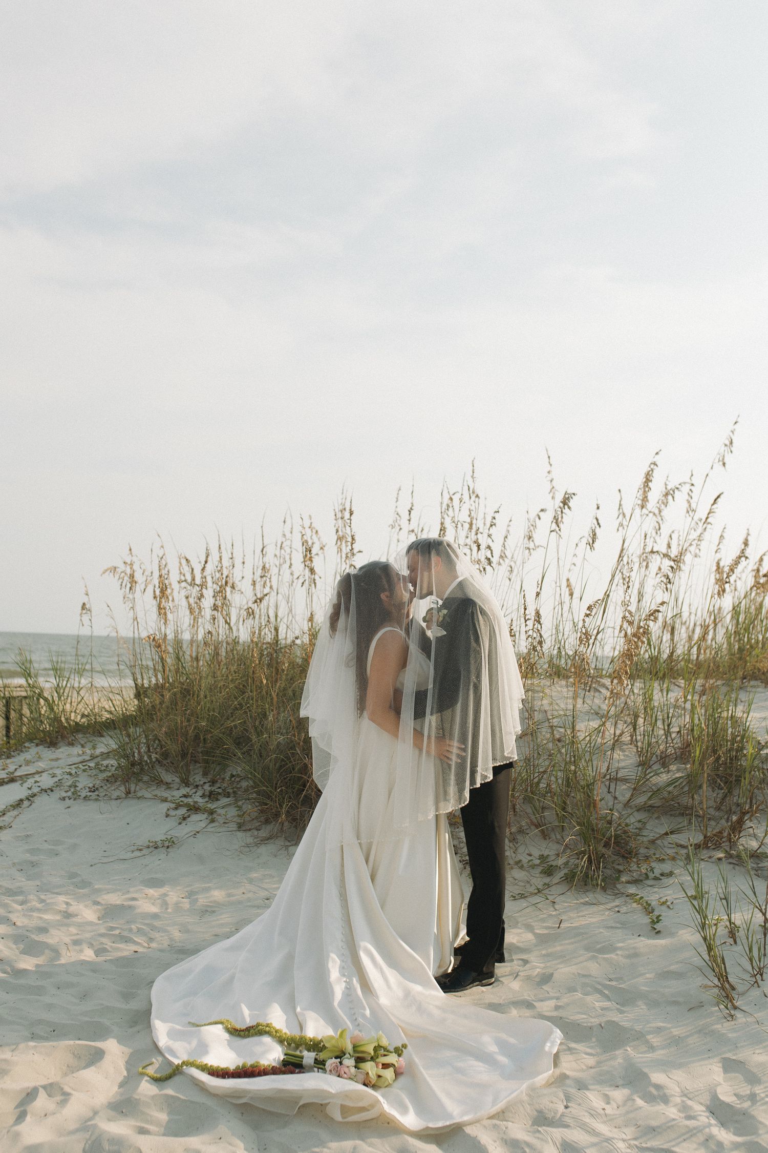 Bride and groom kissing under veil on sandy beach with sea oats, wedding dress train spread on ground.