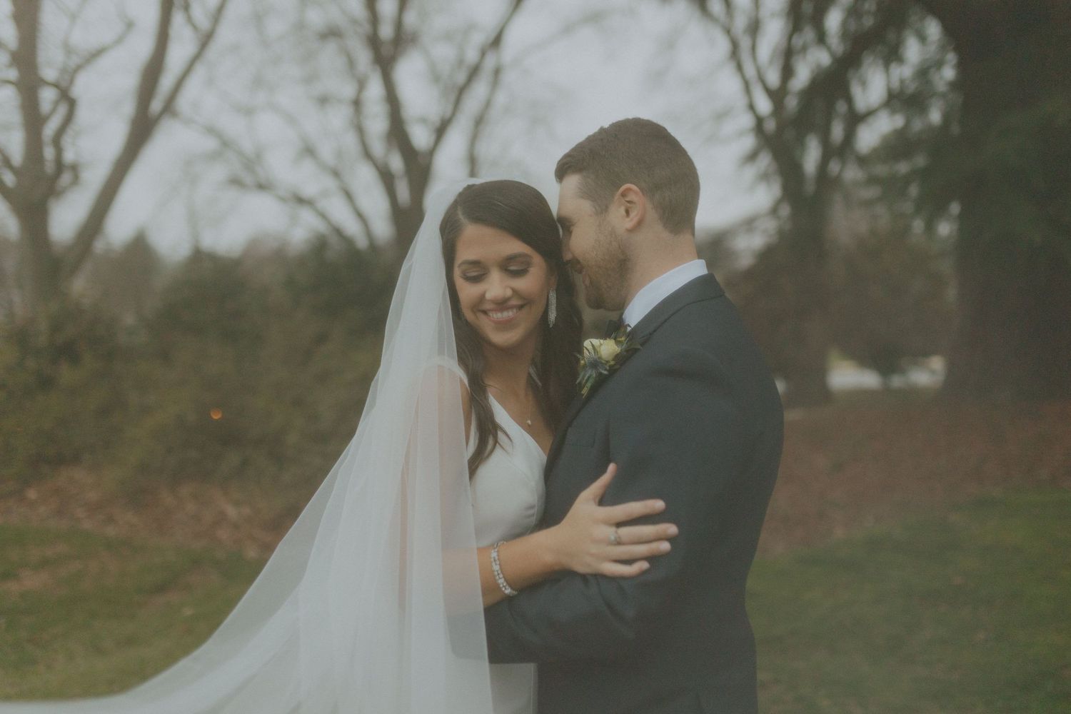 Bride and groom share a tender moment outdoors, with flowing veil and lush trees in the background.