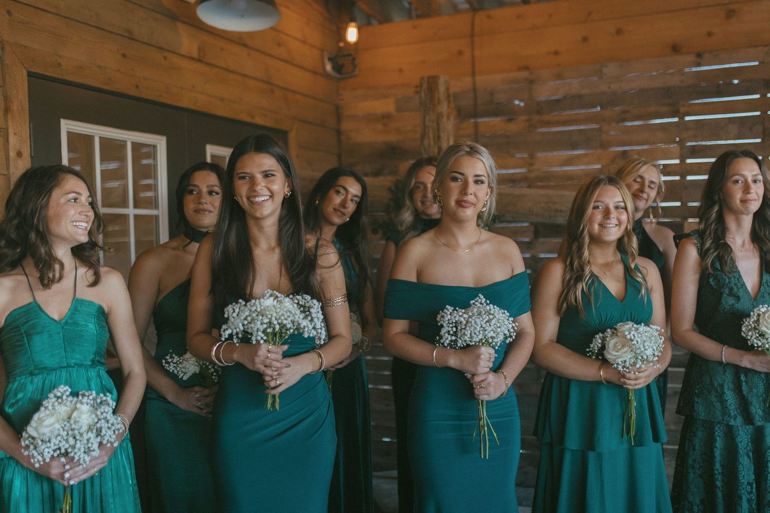 Bridesmaids in teal dresses holding white baby's breath bouquets laughing together in a rustic wooden barn.