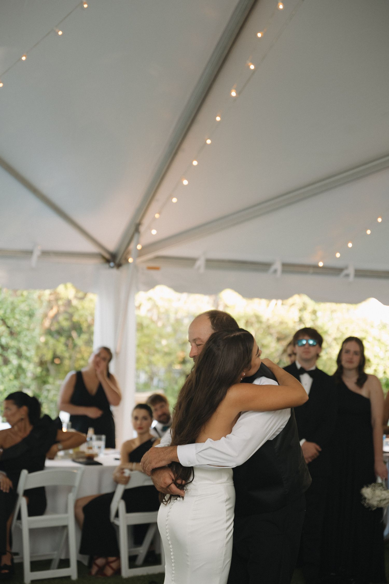 Bride and groom sharing first dance under white tent with string lights at wedding reception.