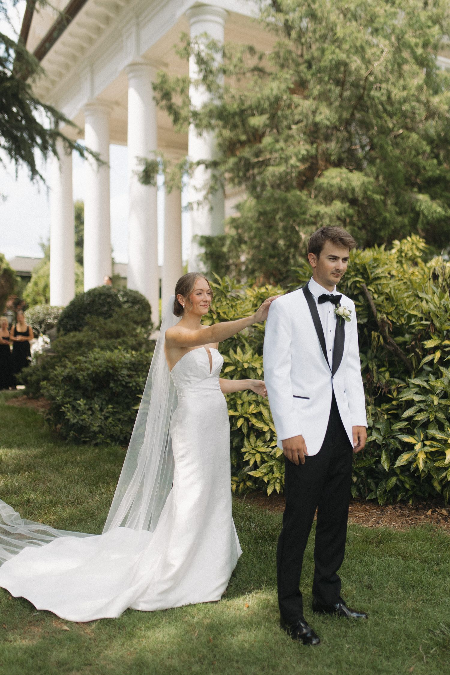 Bride in white gown with veil touches groom in white tuxedo jacket outdoors near pillared building surrounded by greenery.