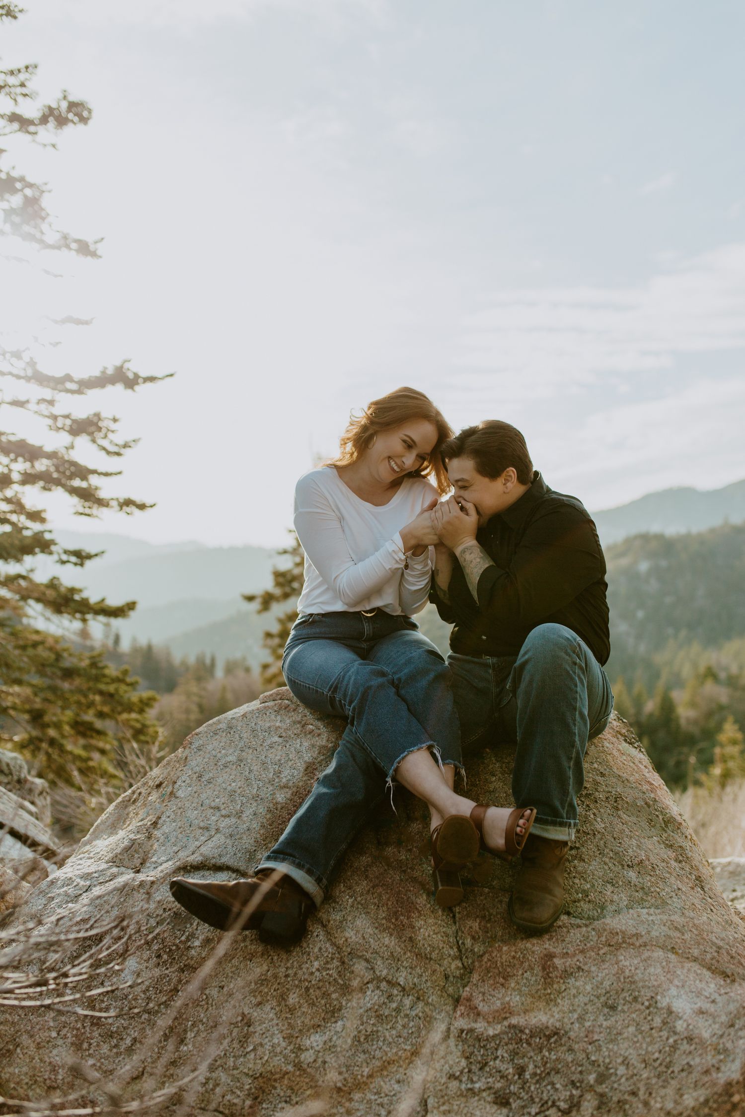 Couple sitting on a boulder sharing a tender moment with mountain forest views behind them.
