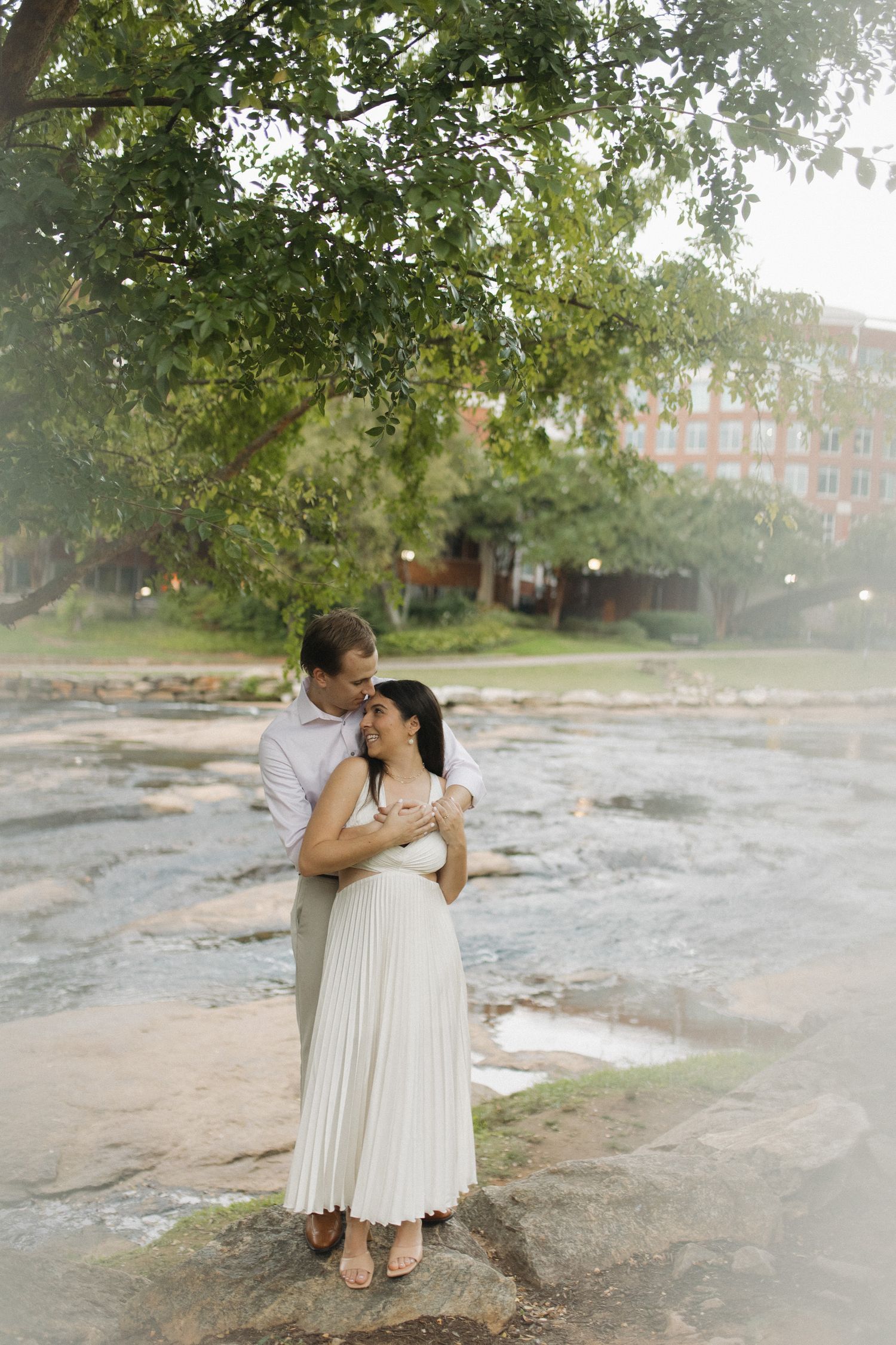 Couple embraces romantically on rocks by a creek, surrounded by lush green trees in soft natural light.