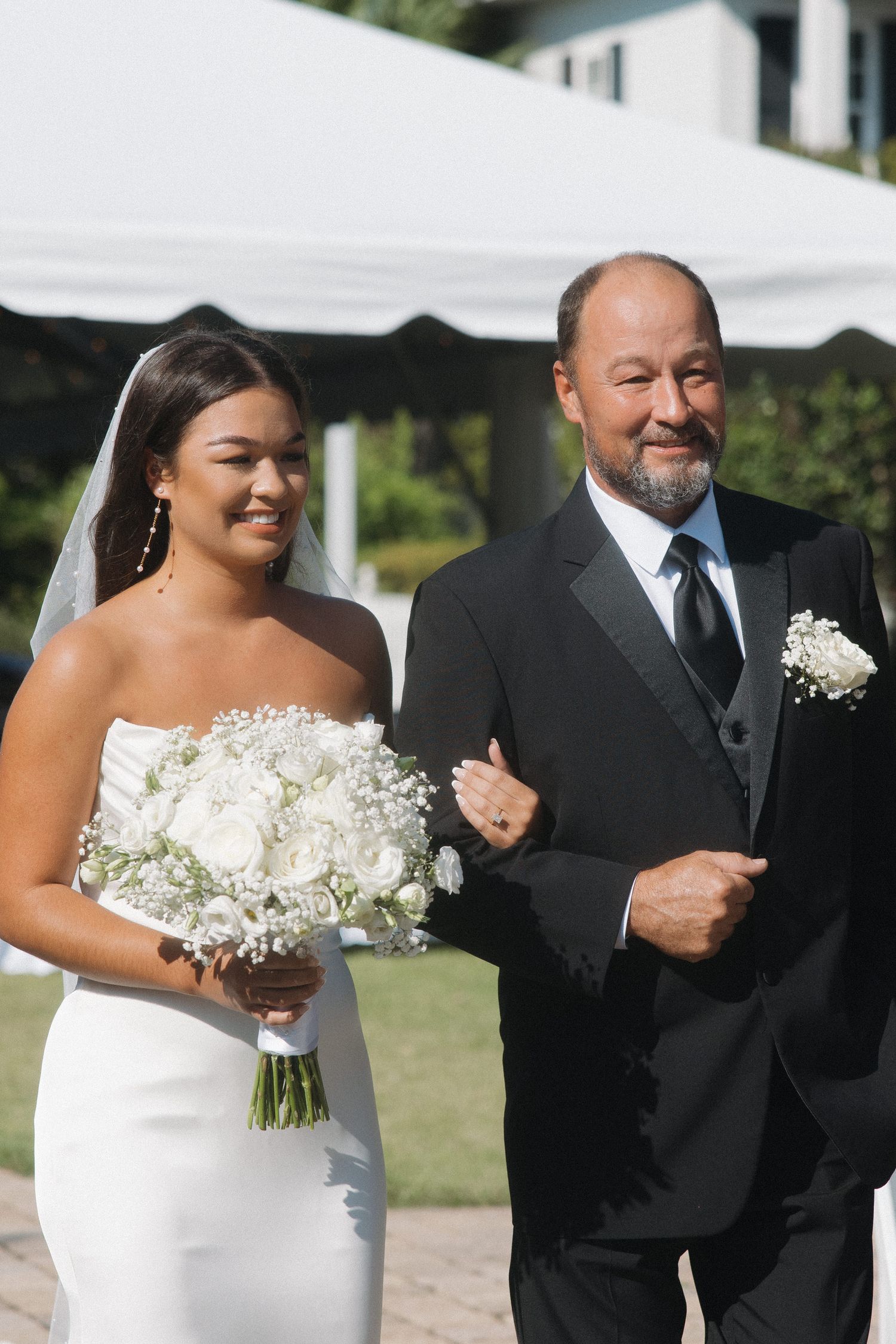Smiling bride in white strapless gown walks down aisle with father holding bouquet.
