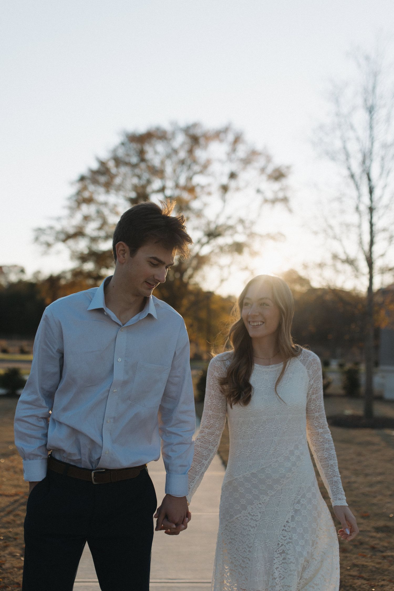 Engaged couple holding hands and walking on a sunlit pathway during autumn sunset.