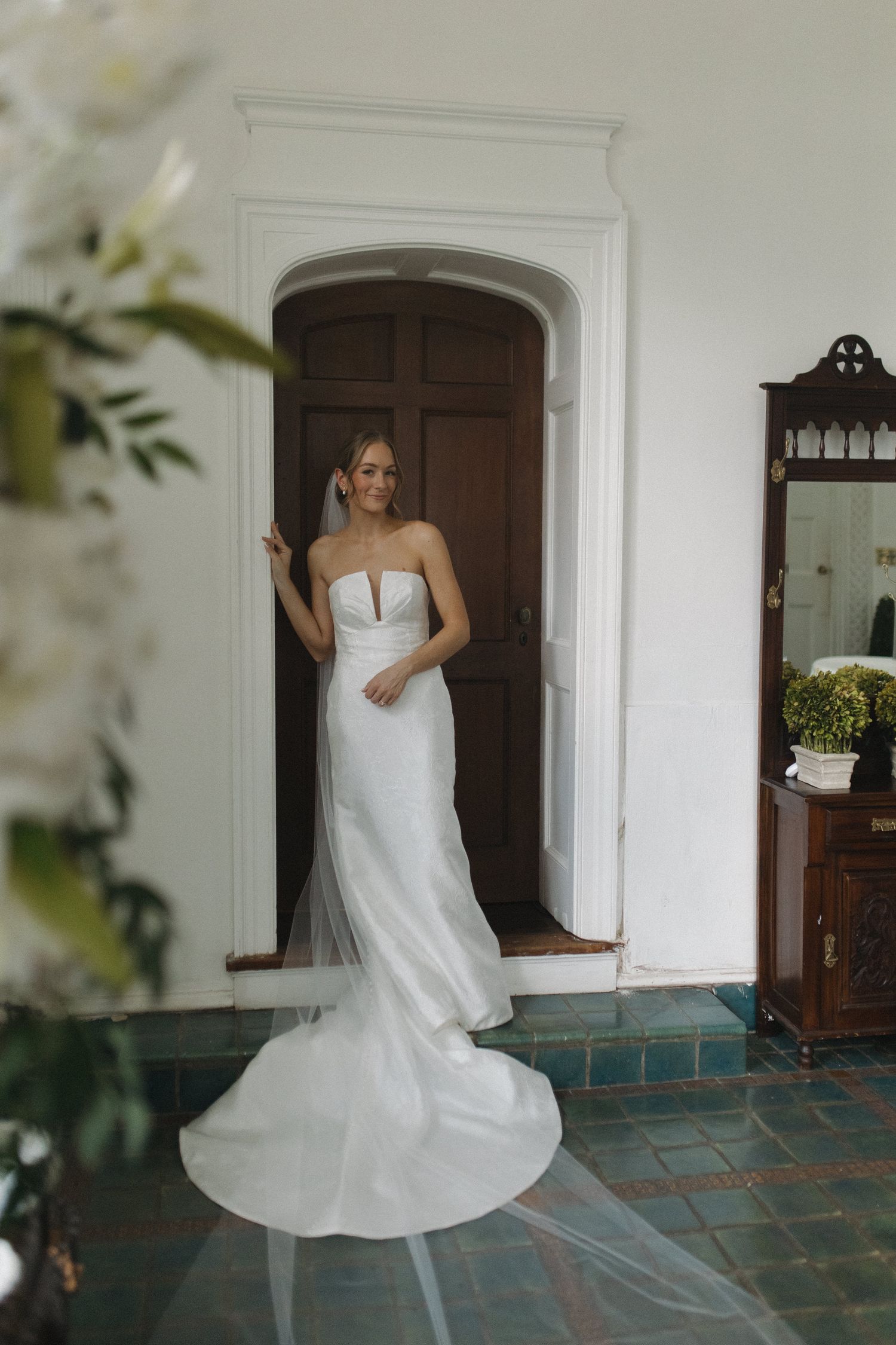 Elegant bride in strapless white gown with veil standing in a doorway of a classic building.