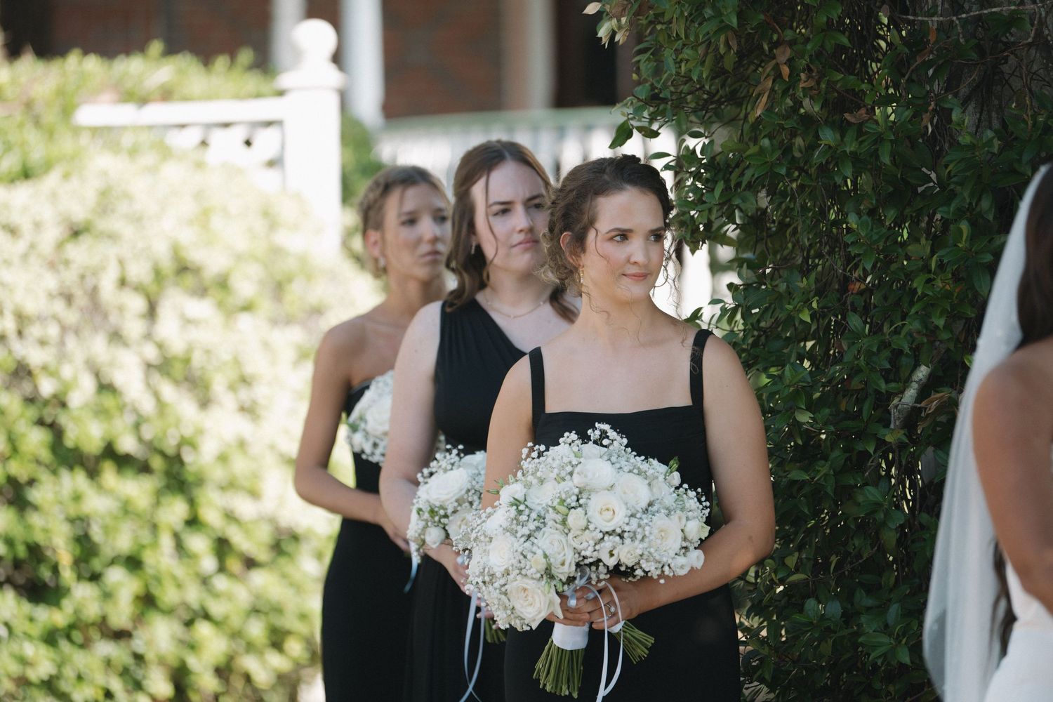 Bridesmaids in black dresses holding white floral bouquets stand outdoors during a wedding ceremony.