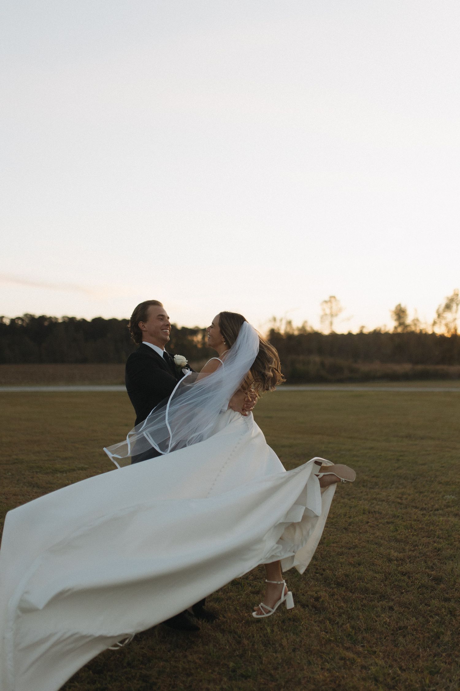 Groom lifts bride in flowing white wedding dress and veil outdoors at sunset on a green field.