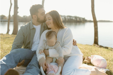 A loving couple sits on a blanket by a lake, sharing a tender moment with their baby in golden sunlight.