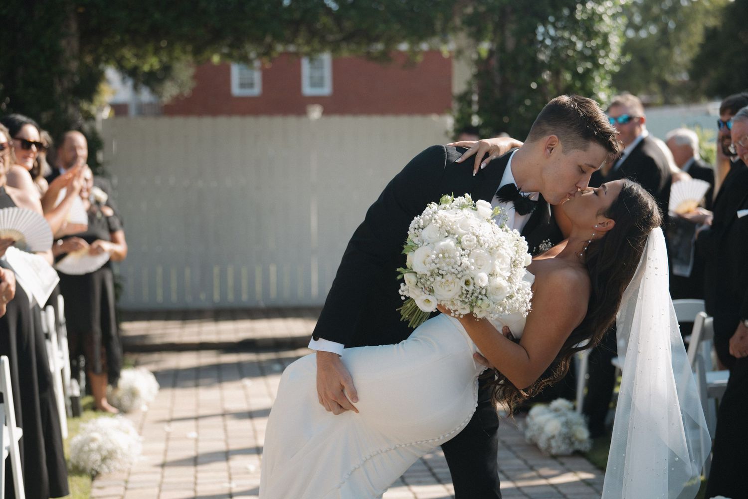 Groom dips bride for a romantic kiss at outdoor wedding ceremony, bride holding white floral bouquet.
