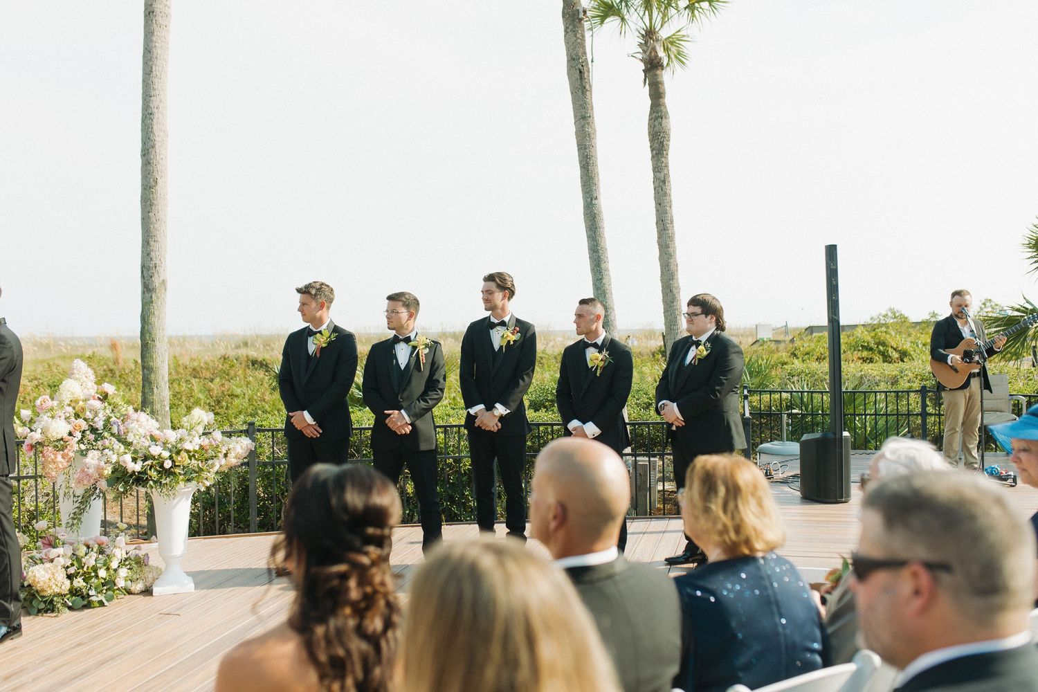 Groomsmen in black tuxedos with boutonnieres stand at an outdoor beach wedding ceremony on a wooden deck with palm trees.