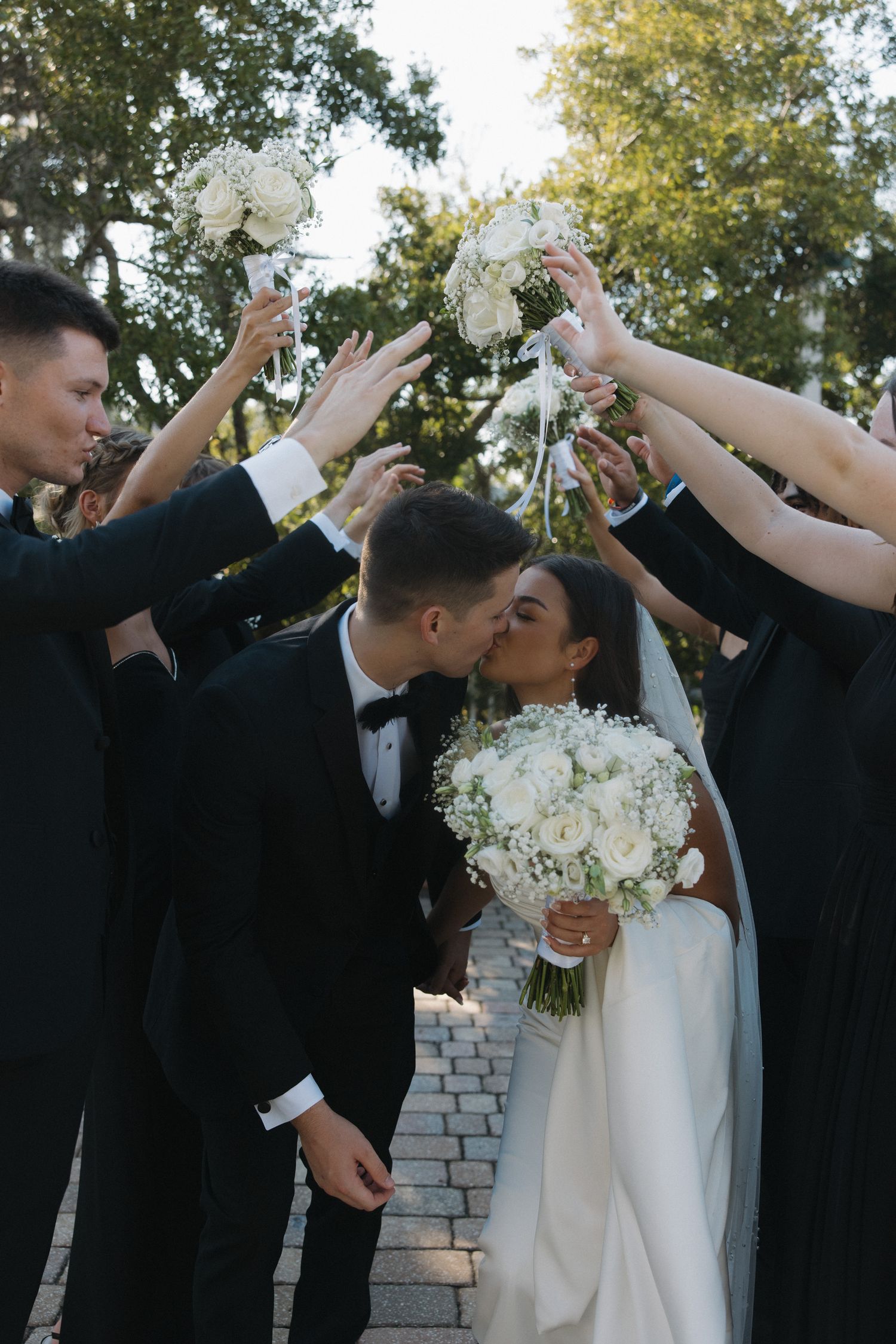 Bride and groom kissing under arch of raised white bouquets held by wedding party on outdoor brick pathway.