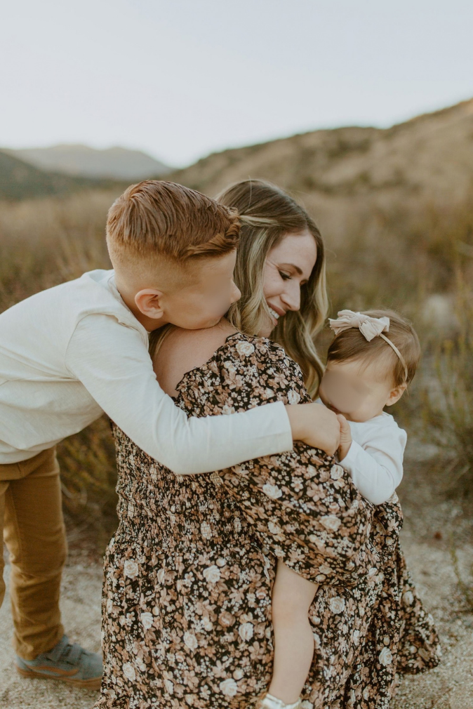 A young boy kisses a smiling woman in a floral dress holding a baby girl outdoors in a desert landscape.