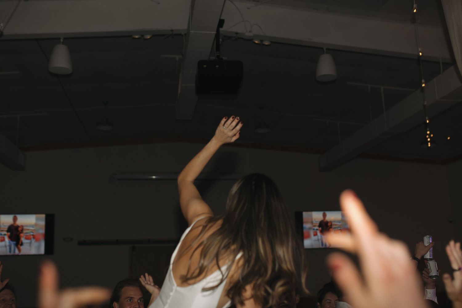 Woman in white dress raises arm at lively indoor party with screens and crowd in background.