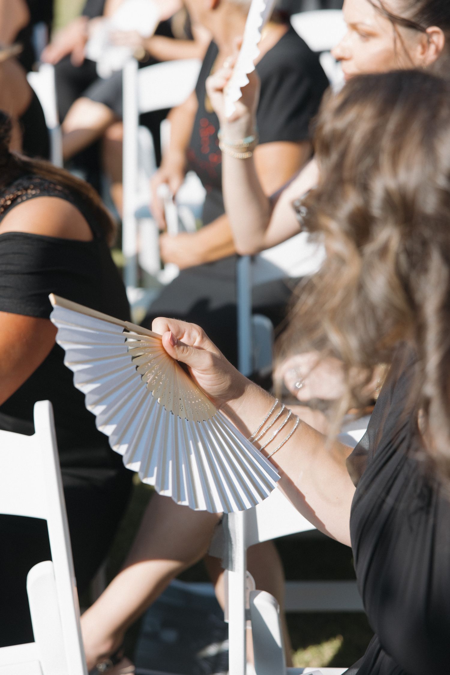 Wedding guests in black dresses using paper fans to stay cool during outdoor ceremony.
