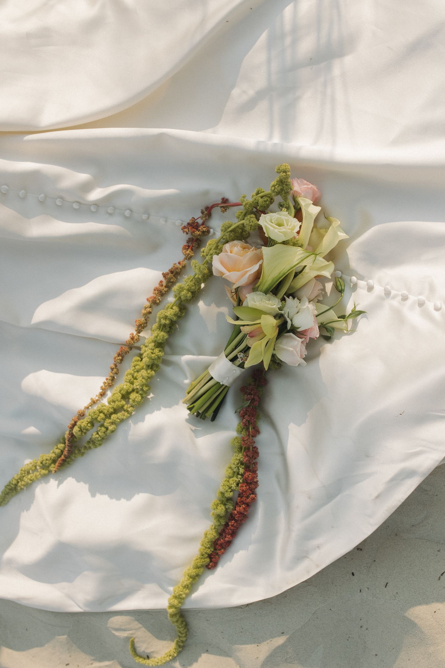 Bridal bouquet of roses and calla lilies with trailing amaranthus on white wedding dress fabric.