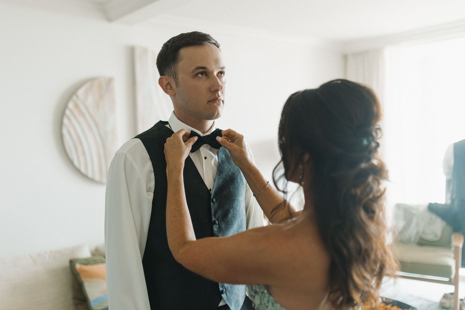 A woman adjusts a groom's bow tie as he prepares for his wedding day indoors.