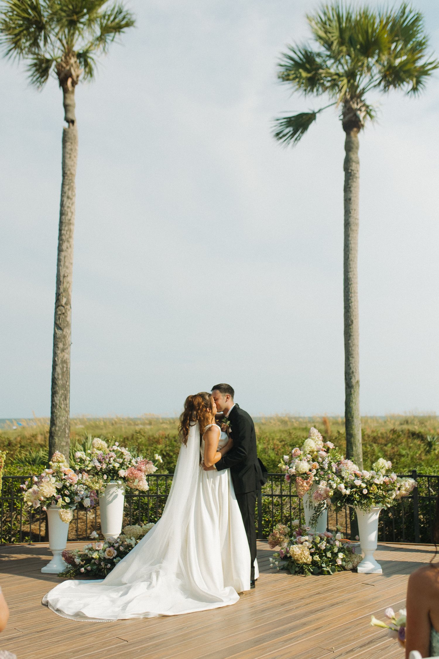 Bride and groom share first kiss at outdoor wedding ceremony with palm trees and floral arrangements by the ocean.