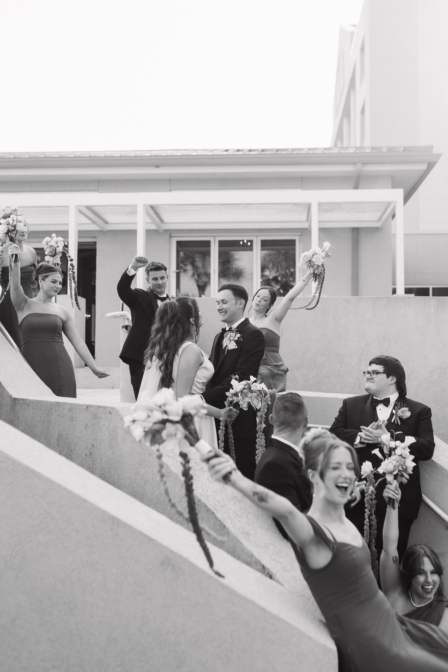 Black and white photo of a joyful wedding party celebrating on outdoor steps, holding bouquets and raising glasses.