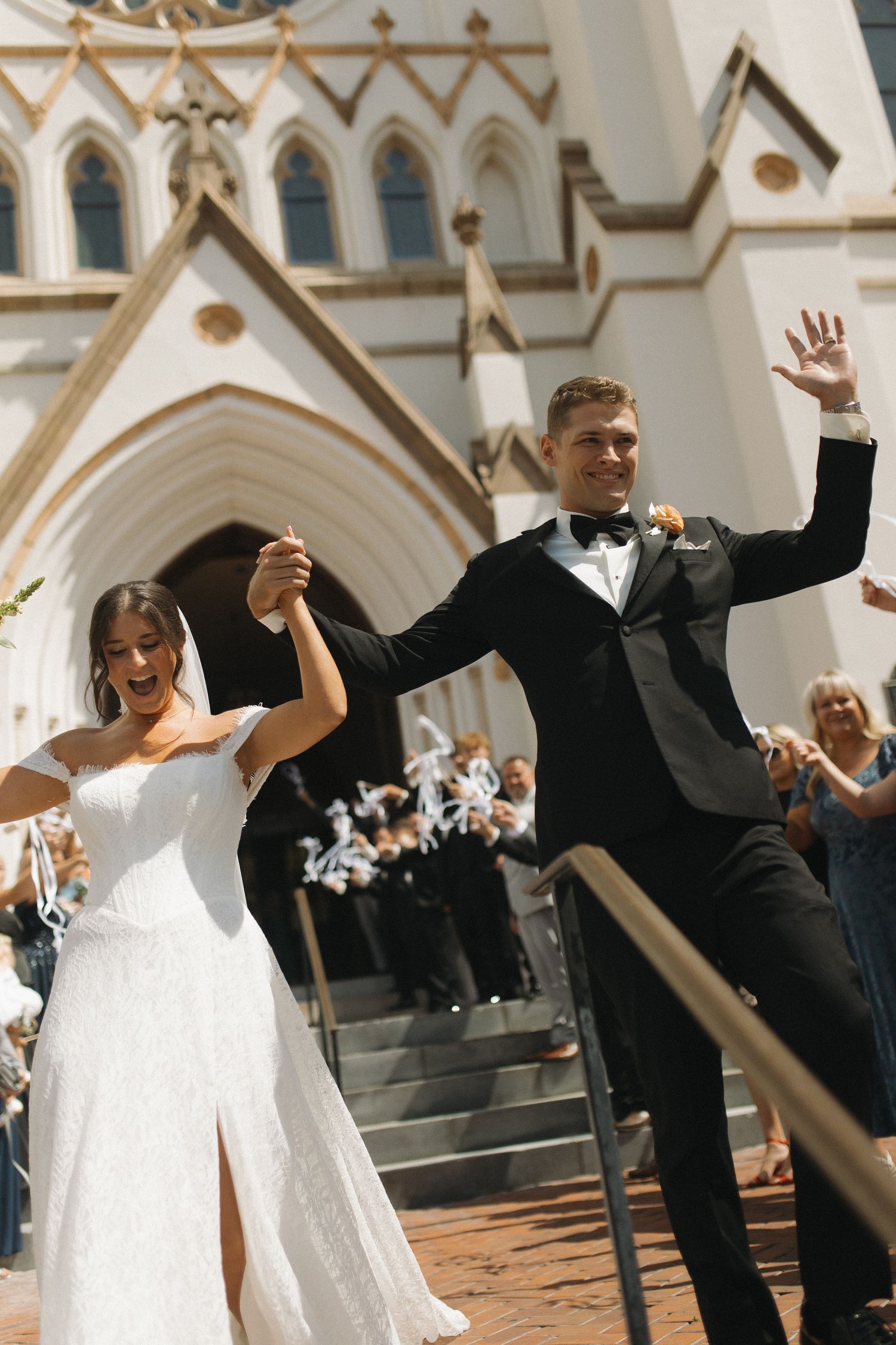Joyful newlyweds exit a grand Gothic church, bride in white gown and groom in black tuxedo, waving to cheering guests.