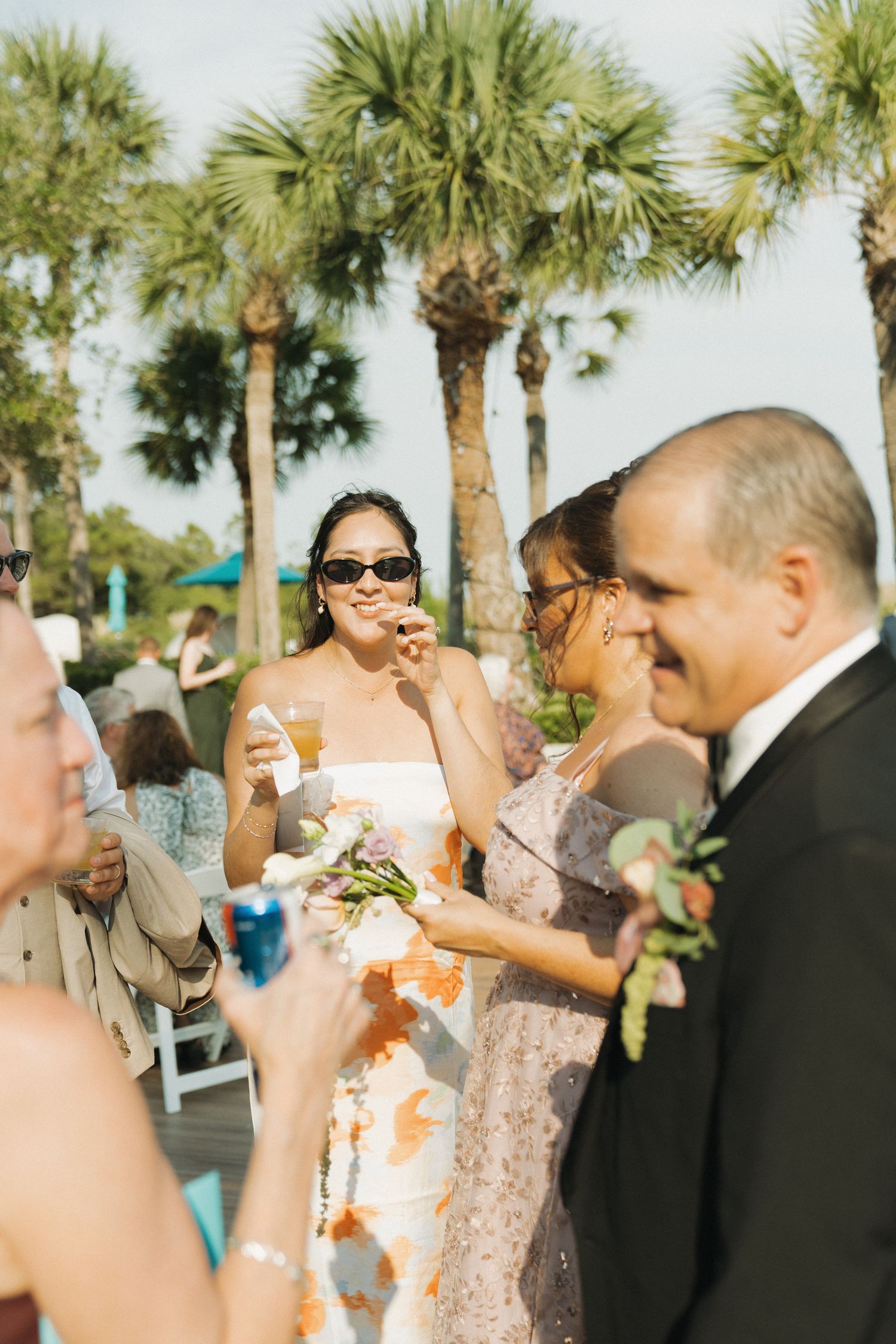 Bride in floral dress with sunglasses holds bouquet at outdoor wedding reception with palm trees.