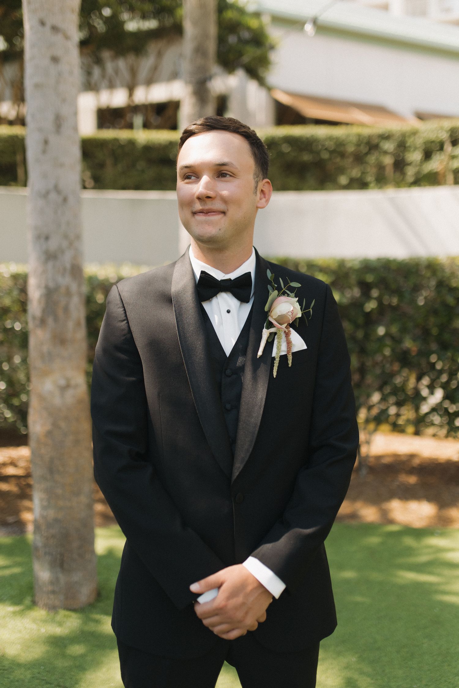 A handsome groom in a black tuxedo with a floral boutonniere smiles outdoors on his wedding day.