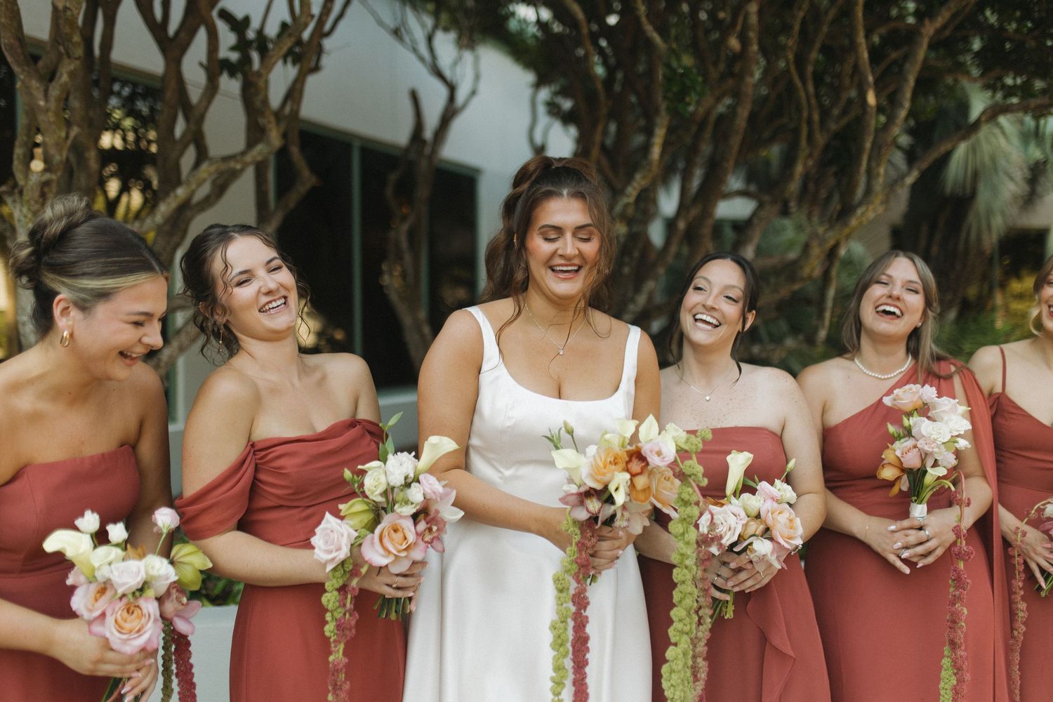 Bride in white gown laughing with four bridesmaids in rust-red dresses holding floral bouquets outdoors.