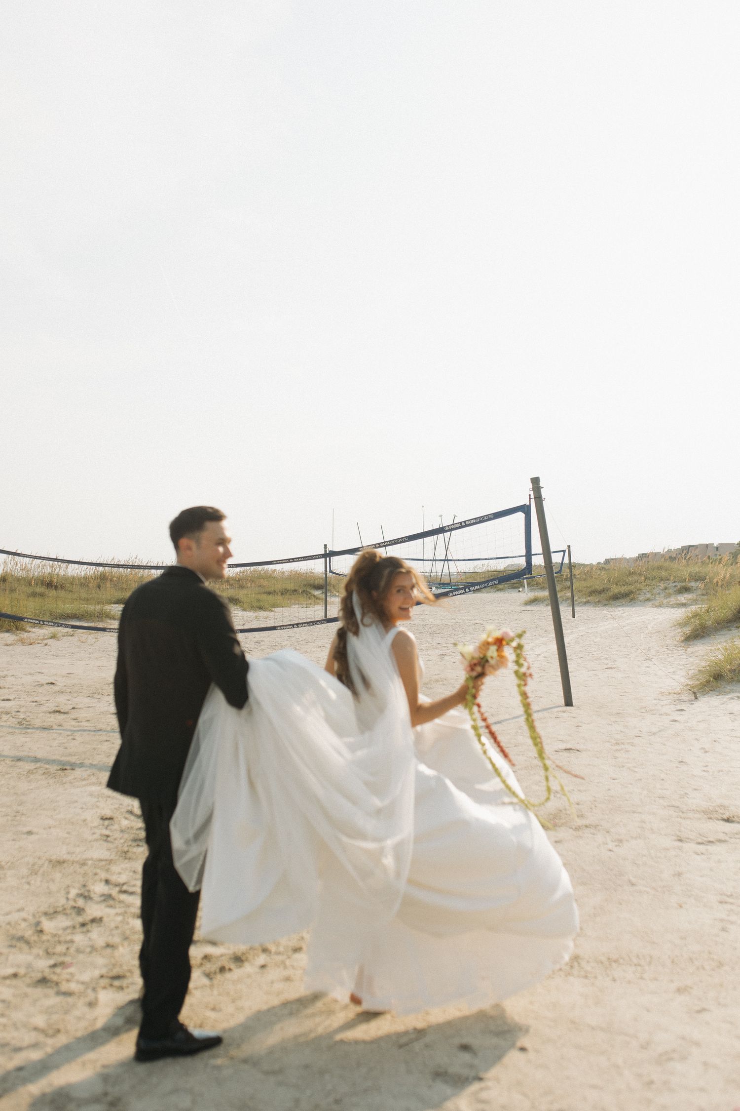 Bride and groom walking on sandy beach, bride holding colorful bouquet in flowing white dress, groom in black suit.