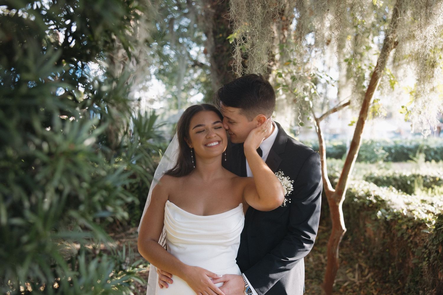 Bride and groom share a romantic moment outdoors under moss-draped trees, groom kissing bride's cheek as she smiles.
