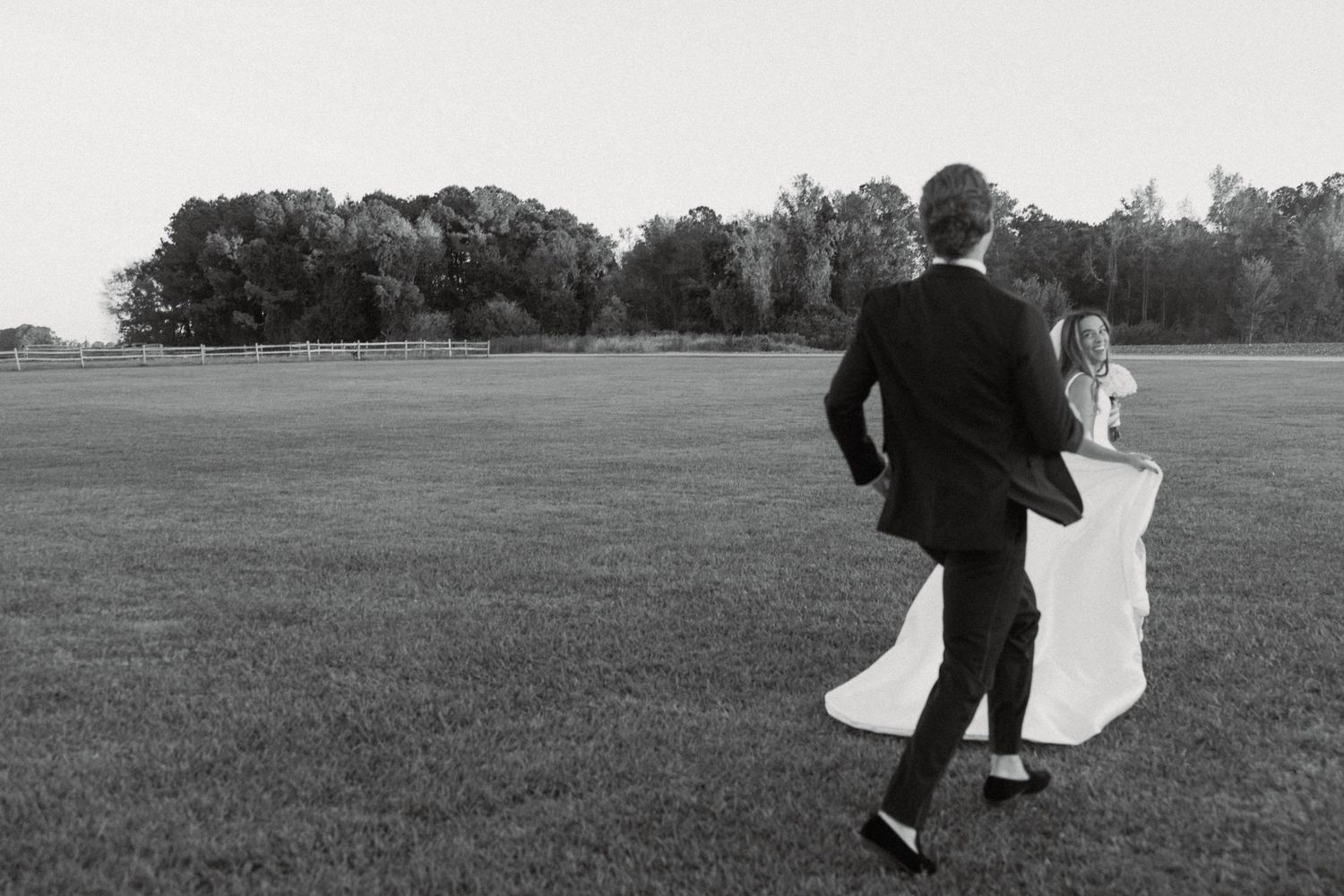 Black and white photo of a bride and groom walking hand in hand across a lush green field toward a tree line.